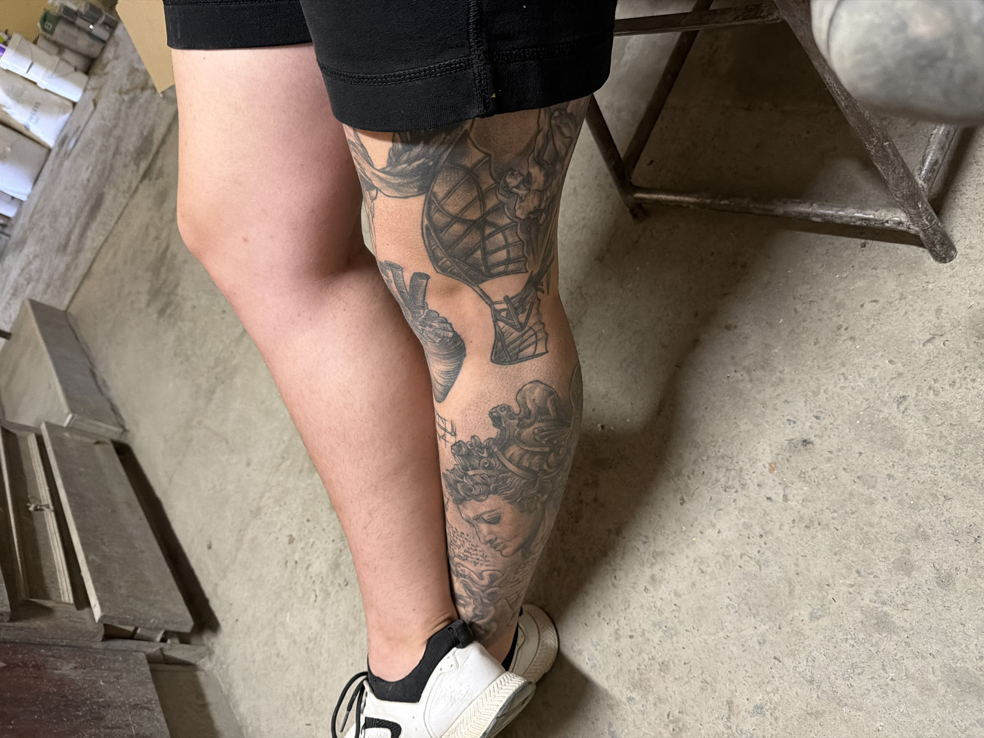An individual's bare legs and a portion of their torso are visible, positioned on a concrete floor. The individual is wearing black shorts and white and black athletic shoes. Their left leg is bent at the knee, with the foot resting on their right thigh. Intricate tattoos adorn the left leg, extending from the ankle to the knee. These tattoos feature classical imagery, including a detailed depiction of a face and what appears to be an early flying machine. The background consists of a concrete floor and a grey, metal structure. The environment suggests an indoor, possibly industrial or workshop setting, based on the presence of a metal frame and the unpolished concrete. No specific location in Chervena Voda, Bulgaria is discernible from the image itself.