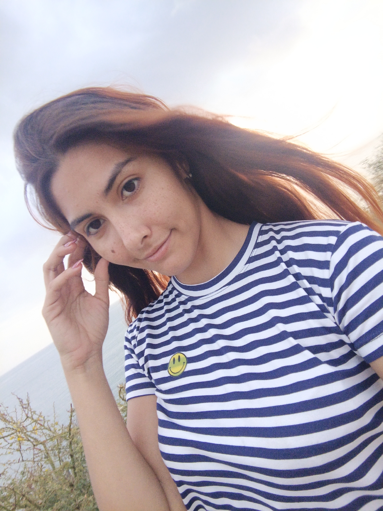 A young woman is the central subject of the photo. She has long, auburn hair that is windblown, brown eyes, and freckles on her face. She is wearing a striped blue and white shirt with a yellow smiley face pin. Her left hand is raised near her face. The background reveals the horizon line of the ocean and sky. There is sparse vegetation in the lower left foreground of the frame. Based on the location information provided, the photo appears to be taken outdoors in Juan Griego, Venezuela.
