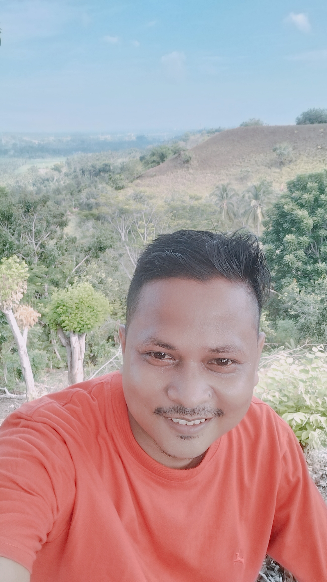The image is a selfie of a man with dark hair and a mustache, wearing an orange t-shirt. He is smiling and looking at the camera. The background is a landscape with trees, hills, and a cloudy sky. The man appears to be in a rural or natural setting, possibly in the outskirts of Lhokseumawe, Indonesia, given the lush greenery and hilly terrain which are characteristic of some areas around the city.