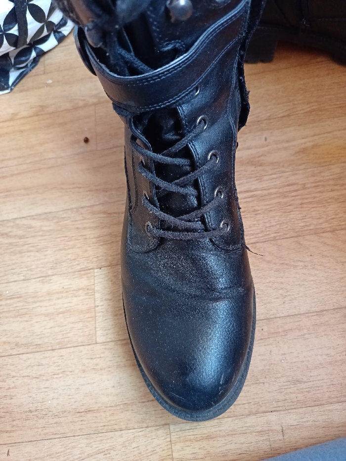 A single black leather combat boot stands upright on a light brown wooden floor. The boot, exhibiting a slightly textured and worn appearance, features black laces threaded through eight pairs of silver-colored metal eyelets. A black leather strap with a metallic, possibly silver-colored, stud or buckle secures the upper shaft. The sole is thick and dark. Scattered dust and a small, reddish-brown speck are visible on the wooden floorboards, which display a distinct grain pattern. In the upper left background, a piece of white fabric with a black geometric pattern is partially visible. No individuals, actions, or direct interactions are depicted. The scene is located in Kamenz - Kamjenc, Germany.