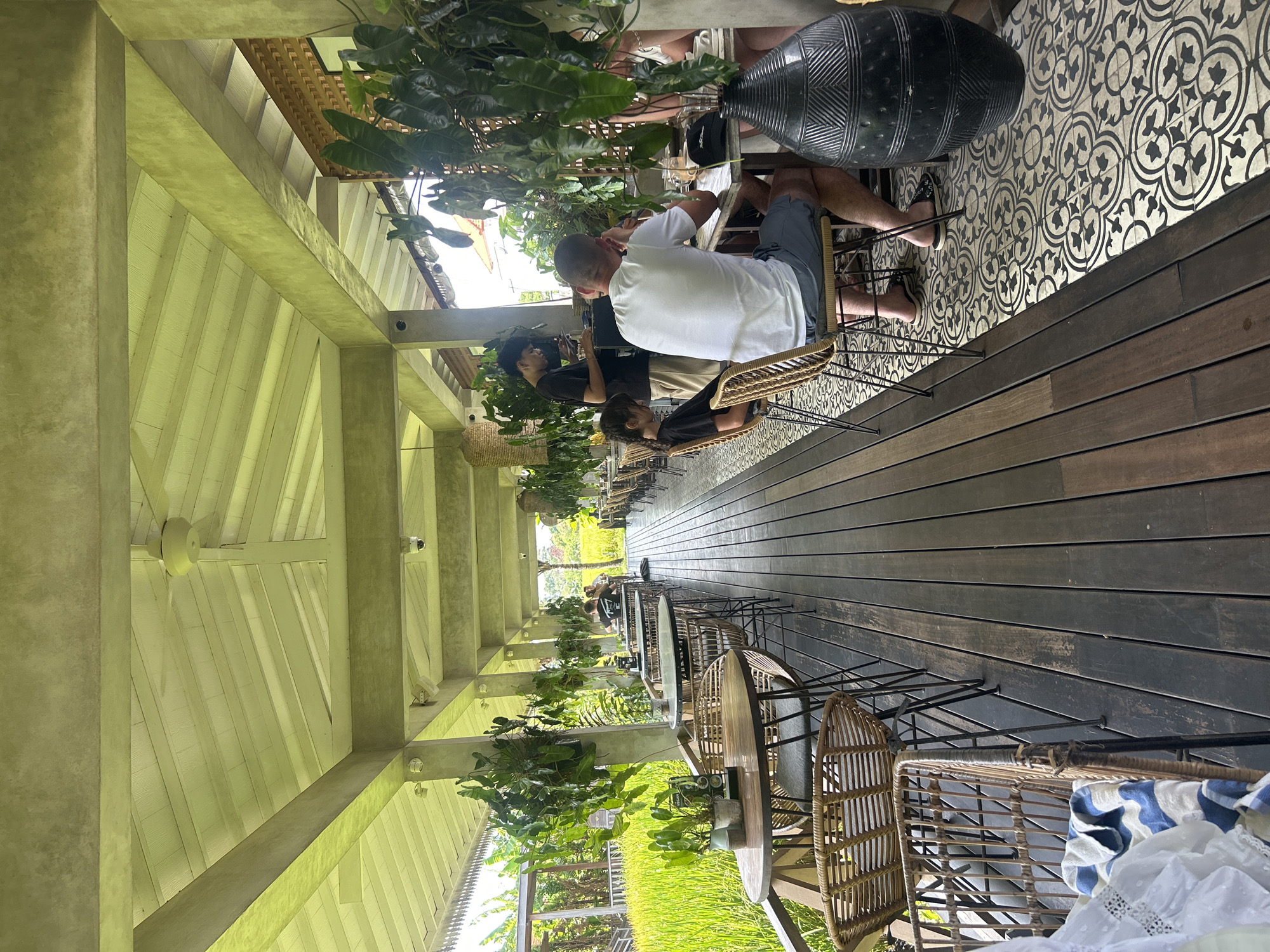 This image depicts the exterior seating area of a restaurant or cafe in North Kuta, Indonesia. The area is covered by a sloped, white ceiling supported by concrete pillars and beams, with a ceiling fan visible in the distance. Hanging plants add greenery to the space.
The seating area features dark wooden decking that transitions to a tiled floor with a black and white floral pattern. Wicker chairs and small, round tables are arranged along the walkway, some with potted plants on them. Beyond the seating area, a lush green rice paddy is visible, suggesting a rural or semi-rural setting within North Kuta.
In the foreground, several wicker chairs and tables are empty. Further down the walkway, a few tables are occupied by diners. One table has a man in a white t-shirt and blue shorts sitting with his back to the camera, looking at a menu or his phone. A young girl in a black t-shirt is seated next to him. Another person, a waiter wearing a black t-shirt, stands at a table, interacting with a diner who is partially visible. Another table further down has diners. A large, dark, decorative vase stands next to the tiled floor.
The scene suggests a casual dining environment, likely designed to take advantage of the natural surroundings. The lighting is natural, indicating it is daytime.
