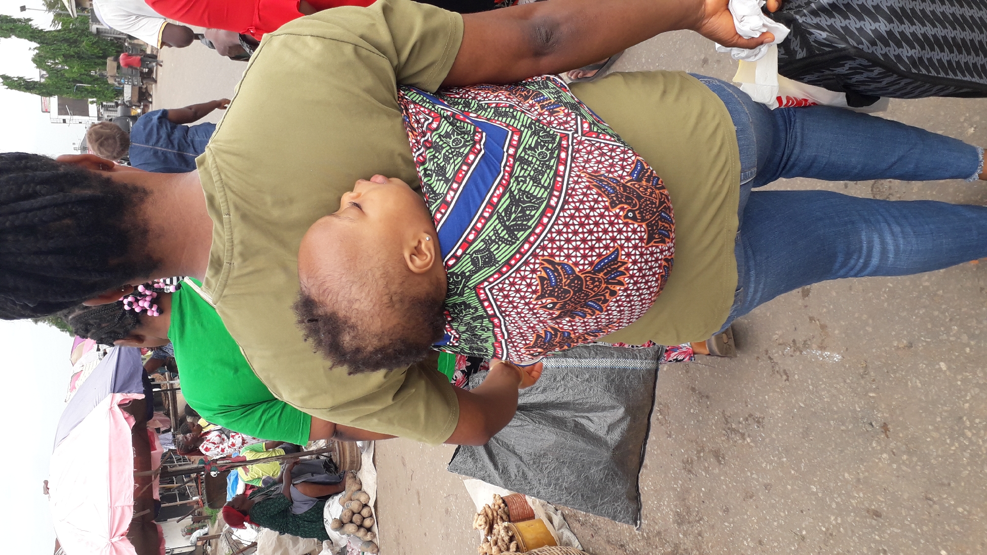 The image shows a woman in a bustling outdoor market, likely in Onitsha, Nigeria. She is wearing an olive-green t-shirt and blue jeans, with a baby secured to her back in a patterned baby carrier. The baby appears to be asleep. The market is filled with vendors and shoppers, with visible stalls displaying items like potatoes, ginger, and other goods. Umbrellas provide shade over some of the stalls. Other people are visible in the background, some in bright-colored clothing. The ground appears to be paved with some debris.