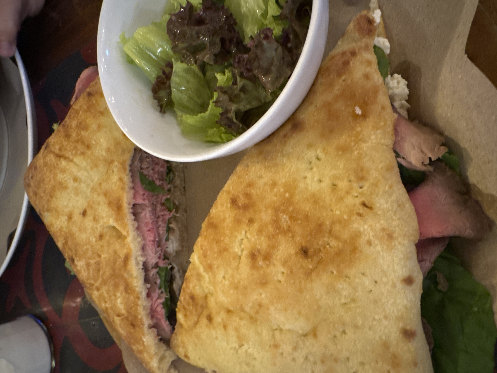 The image shows a close-up of a meal featuring a triangularly-cut sandwich with visible layers of thinly sliced meat (likely roast beef), greenery (possibly spinach or arugula), and a creamy white spread. The sandwich is made with flatbread. Next to the sandwich is a small white bowl containing a green salad with what appears to be lettuce and darker leafy greens. The meal is presented on a brown paper liner. Part of a white plate is also visible on the side. It appears this image was not taken in Ungasan, Indonesia.