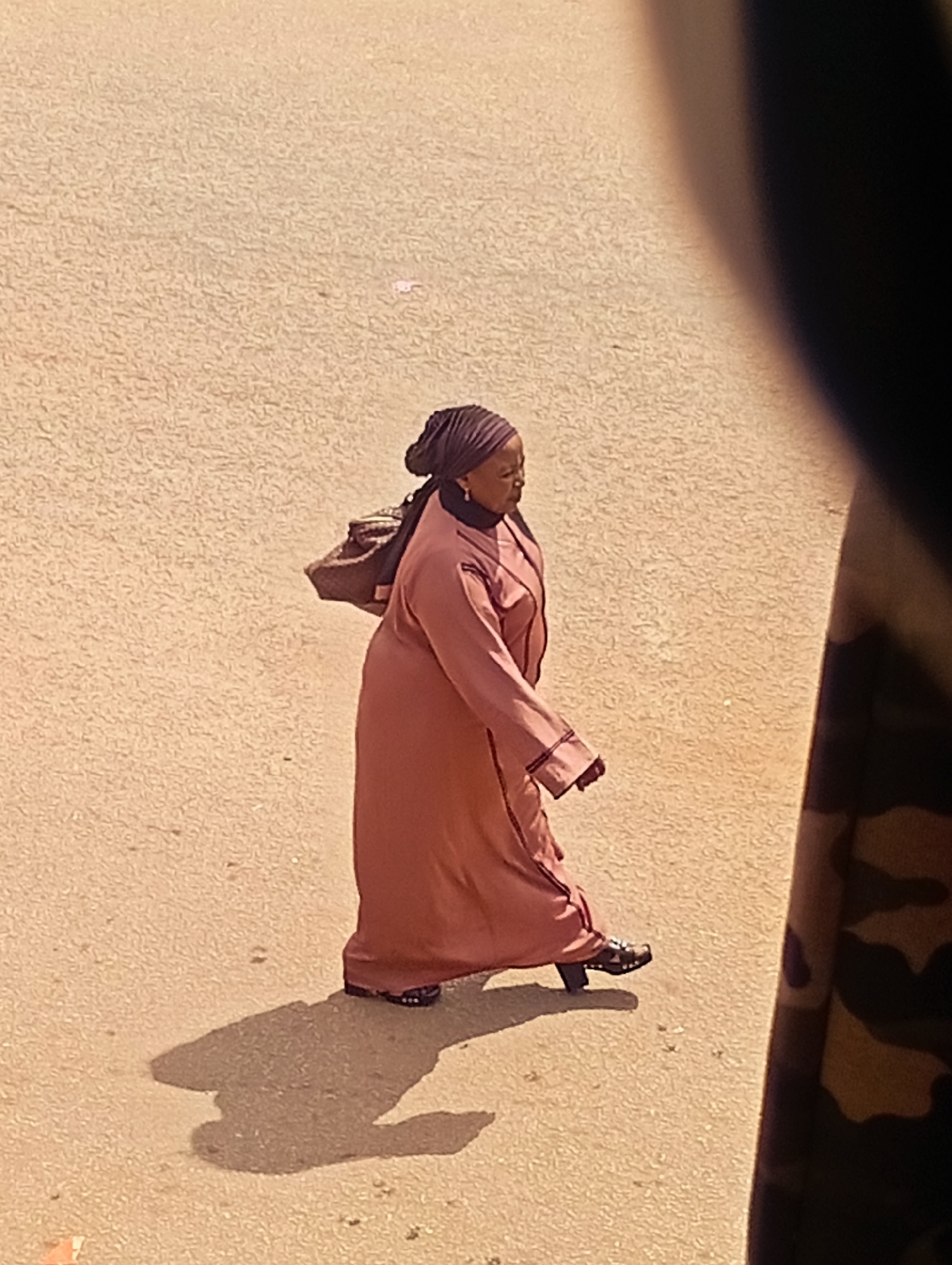 A person, likely a woman, is walking across a paved surface. She is wearing a long, flowing pink garment and a dark headscarf. A brown bag is slung over her left shoulder. She is wearing dark sandals with some embellishments. Her shadow is cast on the ground to her left, indicating the sun is to her right. The ground appears to be a light-colored, textured pavement, possibly asphalt or concrete, with small pebbles and debris scattered across it. The overall lighting suggests it is daytime, likely a sunny day. The scene is outdoors, and the background is a blurred, textured surface. A dark, out-of-focus object intrudes on the right side of the frame, suggesting the photo was taken from behind or to the side of an obstruction. There is no visible text in the image.