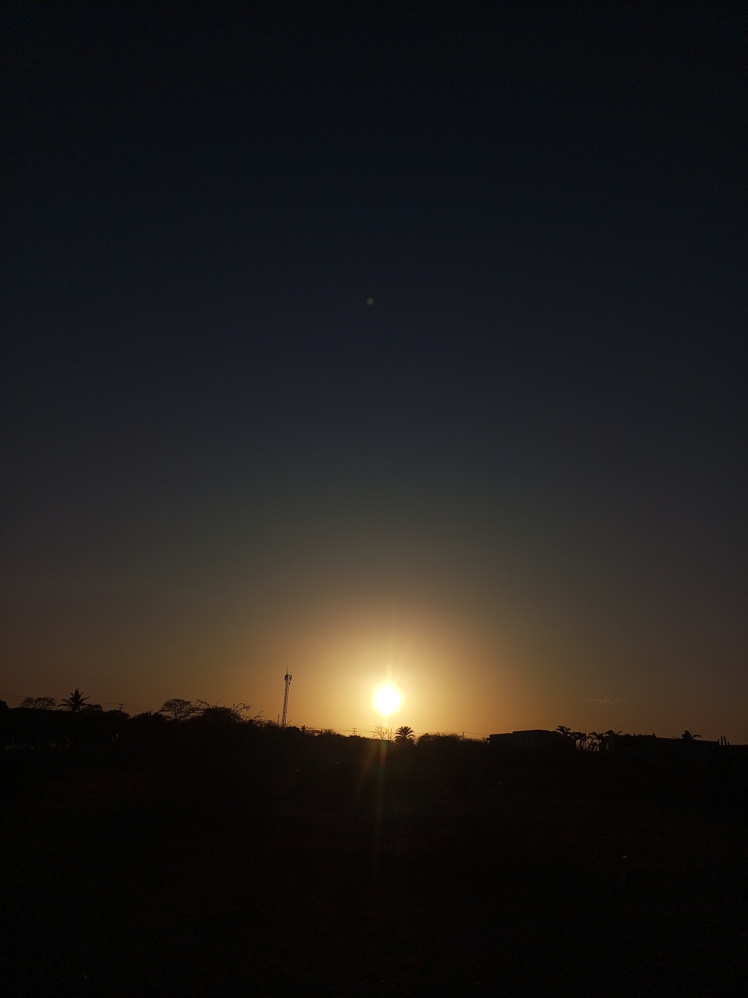 A medium, full outdoor shot displays a sunset over a silhouetted horizon. The sky transitions from a dark navy at the top to a light, golden hue near the horizon where the sun is positioned. Silhouetted against the bright sun are what appear to be trees, buildings, and a communications tower. The foreground is a dark, unidentifiable mass. This image was most likely taken in Juan Griego, Venezuela, given the request.
