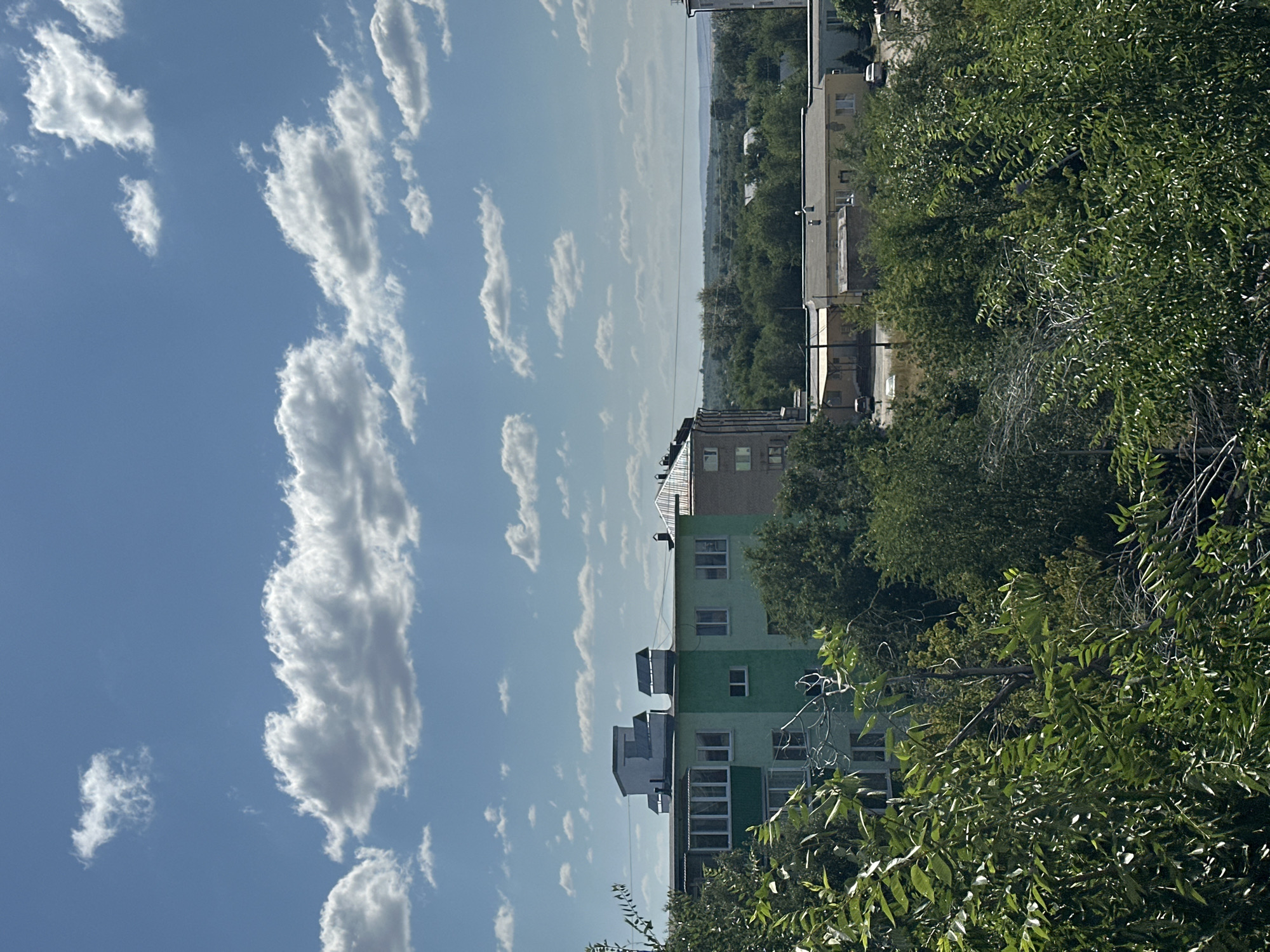 The image depicts a view of the city of Energetik, Russia, on a partly cloudy day. In the foreground, lush green trees and foliage partially obscure the view of buildings and the distant landscape. A multi-story building, painted light green and with a dark green balcony area, is visible in the mid-ground. This building has several windows and a flat roof with some rooftop structures. Behind this building, other structures of varying heights and colors, including a yellow building with a gray roof, are visible, surrounded by dense green trees. The background shows a hazy, distant landscape with more trees and the suggestion of rolling hills under a bright blue sky dotted with white clouds. The overall impression is of a residential area within a natural setting.