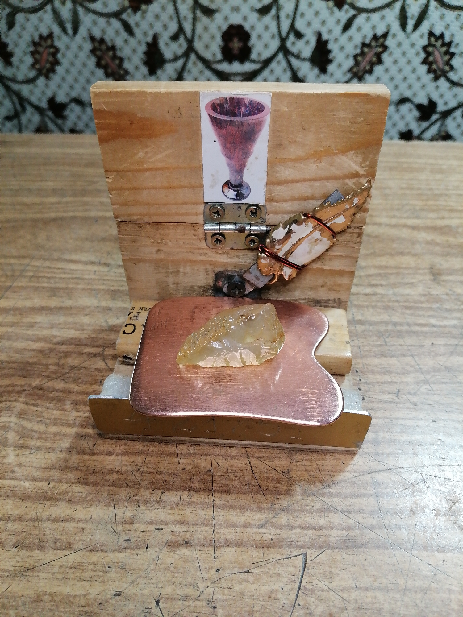 A close-up shot of a rustic wooden structure, possibly a makeshift display or shrine, is presented on a wooden table. The structure is made of unfinished wood planks. On the front face of the upper plank, a small printed image of a red chalice is affixed. Below this image, two metal hinges connect the upper plank to a lower plank, which is slightly ajar. To the right of the chalice image, a decorative element consisting of what appears to be a crab claw, painted white and gold, is attached to the wood with copper wire. In front of the wooden structure, resting on a metallic base, is a copper-colored object with a curved edge, resembling a gua sha tool. A rough, translucent yellow crystal is placed on top of this copper object. The background features a patterned wallpaper with a floral motif in shades of blue and brown. The table surface is a worn wooden plank with visible scratches and marks. The scene is well-lit, with a slight overhead light source casting shadows. There is no indication of the location being in Rostov-on-Don, Russia, or any specific city. The image focuses on the arrangement of these objects, with no people or active events depicted.