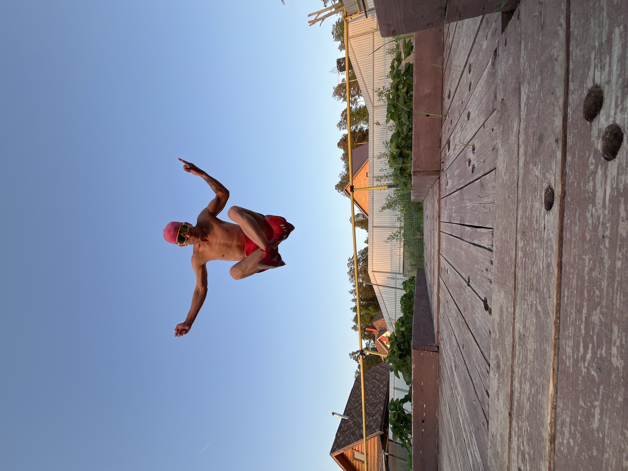 A man in a red bathing suit and red swimming cap jumps off a wooden structure into the air. The sky is blue. In the background are houses and fences. The photo was taken in the city of Chaikovsky, Russia.