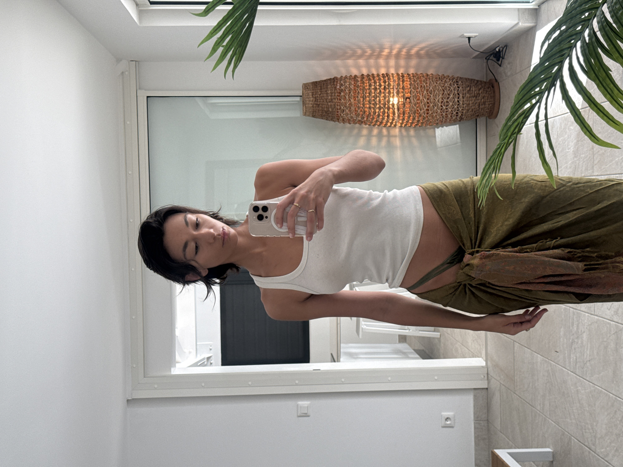 A young woman is captured in a mirror selfie, holding a smartphone with a clear MagSafe case in her right hand, positioned to take the photo. She is standing with her torso slightly turned, looking towards the camera with a neutral, calm expression. She has dark hair, styled short and appearing slightly damp or swept back. She is wearing a white ribbed tank top that exposes her midriff, and an olive green, flowing garment, possibly a wrap skirt or sarong, which is tied or gathered at her hip. Several rings are visible on her fingers.
The setting appears to be a bright, modern interior, possibly a hotel room or residential space, consistent with a location like Saint-Martin, France. The walls are predominantly white, with a light-colored tiled floor. Behind the woman, there is a large window or glass door that reflects parts of the room, including what looks like a white table or counter and a dark, possibly wooden, structure or door frame.
A notable decorative element is a cylindrical wicker light fixture hanging on the wall, casting a patterned shadow. Overhead, green fronds of a plant, likely a palm, extend into the frame from the top left corner. An electrical outlet is visible on the lower white wall. The overall impression is one of a relaxed, sunlit environment. There is no discernible text visible in the image.