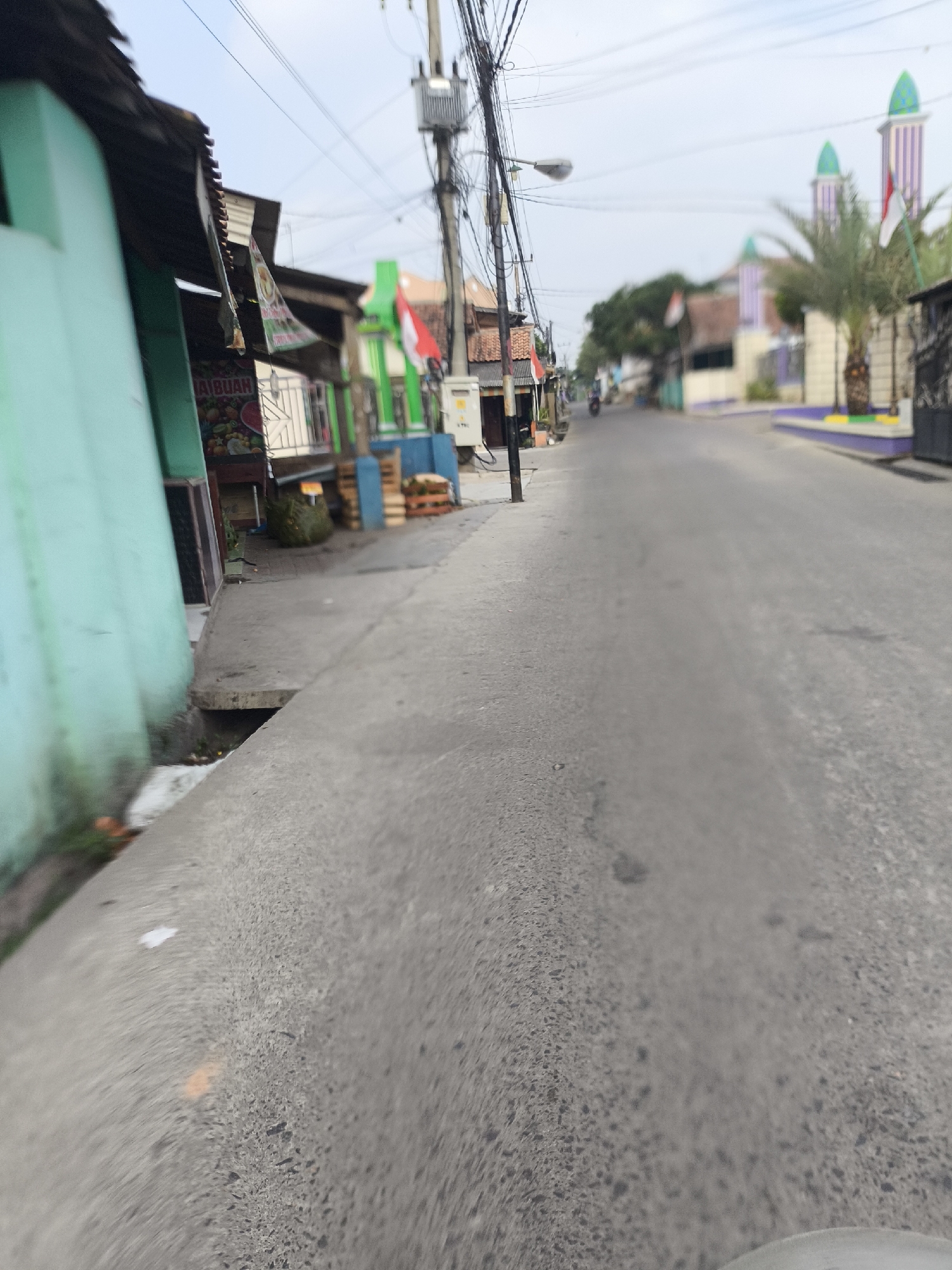 The image depicts a street scene in Cilegon, Indonesia, characterized by a paved road and a sidewalk on the left. The sidewalk features a turquoise-colored building with a corrugated metal roof, possibly a shop or residence. Adjacent to it is a stall or storefront with signage, displaying images of fruits and the word "BUAH" (fruit). Further down the street, more buildings and structures are visible, including what appears to be a mosque with distinctive purple and green striped minarets in the distance. Several Indonesian flags are displayed, suggesting a national holiday or patriotic event. A utility pole with numerous wires stands prominently on the right side of the road. The overall environment appears to be a moderately developed urban or suburban area. The image is taken from a low vantage point, suggesting the photographer may have been on a motorcycle or bicycle. The perspective is slightly distorted, creating a fisheye effect. No individuals are clearly identifiable in the scene, though a motorcycle is faintly visible in the distance on the road. The sky is overcast.