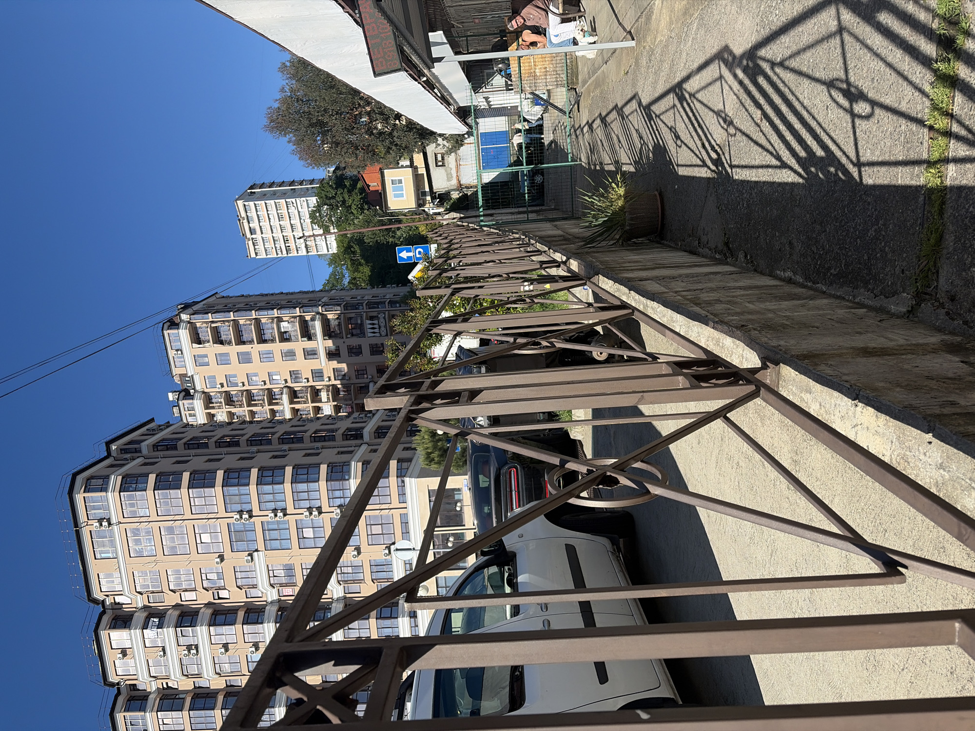 The original image, captured in a vertical orientation, depicts a bright, sunny urban street scene in Sochi, Russia. When viewed horizontally, the immediate foreground is dominated by a sturdy brown metal fence or railing that spans the lower and right side of the frame, casting prominent, angular shadows across a concrete wall and paved ground.
To the left, multiple modern multi-story buildings rise against a clear blue sky. These structures feature numerous windows, balconies, and external air conditioning units, suggesting residential or commercial use. Below these buildings, several cars are parked along the street, including a white van or car and a dark-colored sedan.
On the right side of the street, beyond the fence, a paved sidewalk abuts a low concrete wall. Strong, well-defined shadows from the fence fall onto this wall and the adjacent ground, where patches of green grass are visible. A large potted plant sits near the wall. Further into the midground, a blue traffic sign with two white arrows (one pointing left, one appearing to point downwards or slightly right) is mounted on a pole.
In the background on the right, there's a utility or service area enclosed by a wire fence, containing what appear to be blue and white metal units, possibly for storage or waste. A person is dimly visible in this area, seemingly sitting or engaged in an activity. More buildings and trees are visible in the distance under the bright, cloudless sky. The strong, crisp shadows indicate a clear, sunny day, likely mid-morning or early afternoon. No legible text is visible other than the pictorial traffic sign.