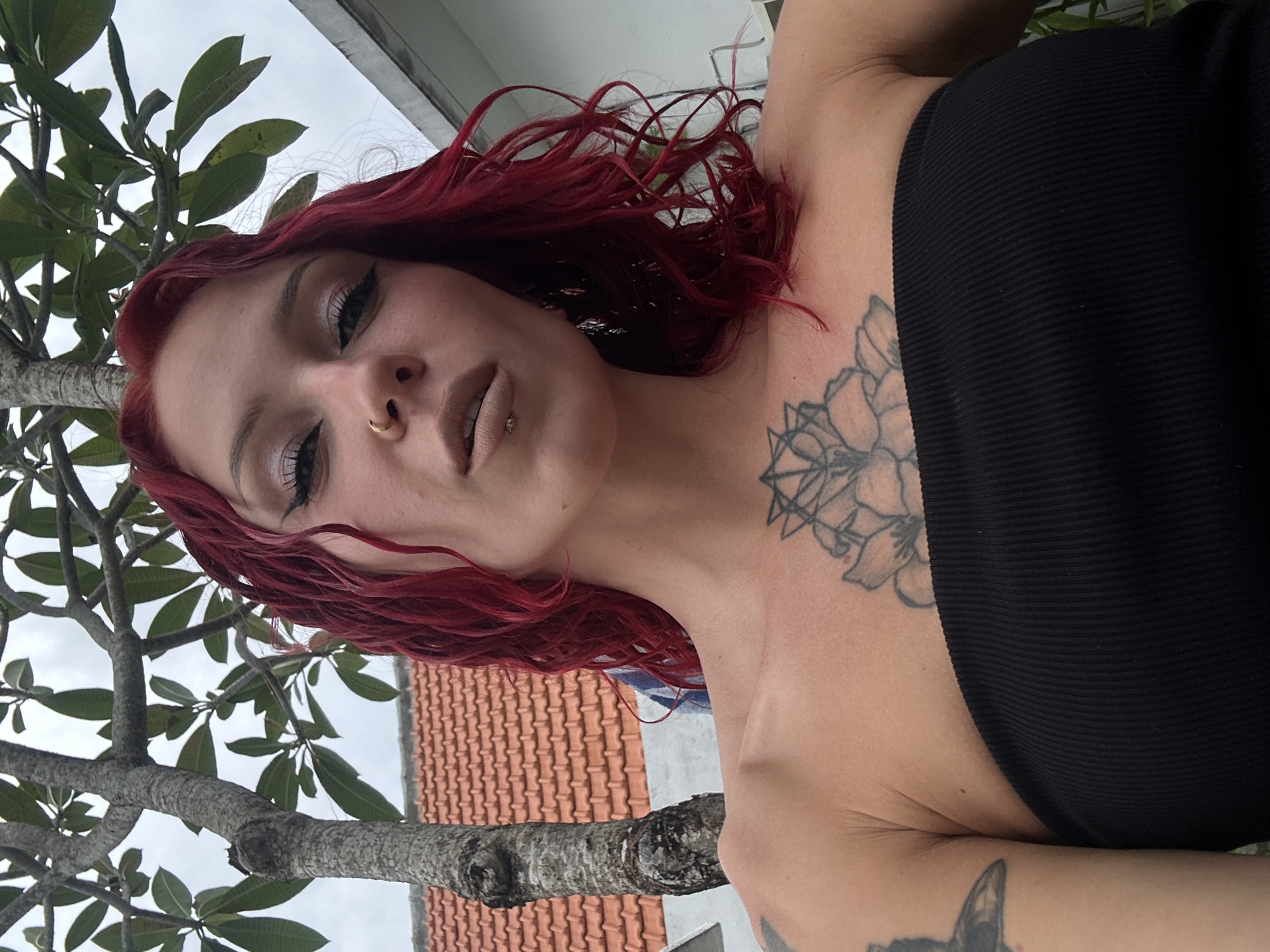 This is an outdoor, low-angle portrait featuring a woman with vibrant red, wavy hair. She has fair skin, distinctive eye makeup with winged eyeliner, and light-toned lipstick. Her facial piercings include a gold hoop in her left nostril and a small stud on her lower lip. She is wearing a black, ribbed, strapless or off-the-shoulder top. Visible tattoos include a prominent floral design on her upper chest and collarbone, and a partial tattoo on her upper right arm, possibly depicting an animal.
The woman is looking directly towards the camera, with her head tilted slightly upwards and her lips gently parted. The scene is set outdoors, with a large tree featuring green leaves and a thick trunk dominating the left and upper portions of the background. Behind the tree, an overcast sky is visible, along with a reddish-orange tiled roof and a light-colored wall of a building, suggesting a residential or urban environment.
The lighting is soft and diffused, indicating an overcast or cloudy daytime. The tropical appearance of the tree and the architectural style of the tiled roof are consistent with the provided location context of North Kuta, Indonesia. The image appears to be a posed photograph. No text is visible in the image.