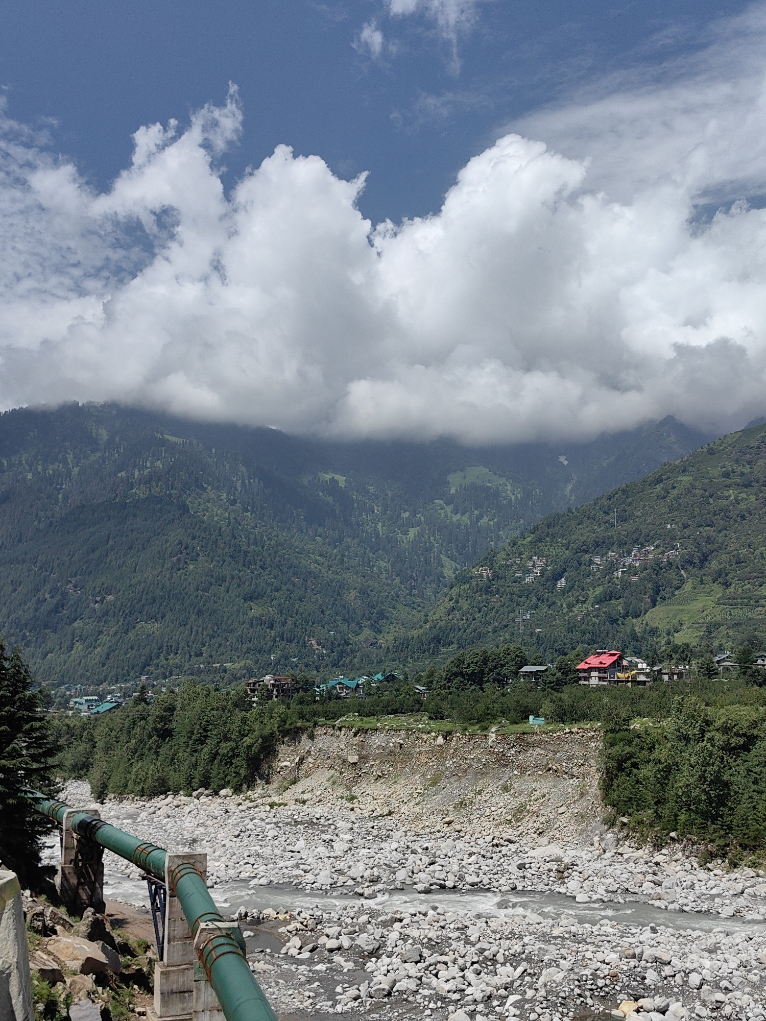 The image shows a wide mountain valley in Palchan, India, under a partly cloudy blue sky. In the foreground, a rocky riverbed is visible with a shallow, rocky river flowing through it. A large green pipeline supported by concrete pillars crosses the river. Trees and dense vegetation line the banks of the river. In the mid-ground, there are scattered houses and buildings, some with red roofs, nestled among more trees. The background is dominated by steep, heavily forested mountains, with the upper slopes of some mountains obscured by clouds. The overall scene depicts a rural mountainous landscape.
