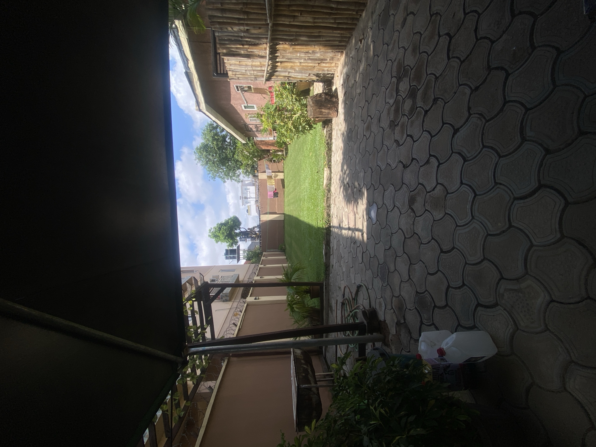 The image depicts a backyard in Barden Yana, Nigeria, viewed from under a dark overhang. The foreground is paved with interlocking grey tiles. To the left, a large, rusted metal basin on a stand is partially visible, along with some potted plants. Two large, translucent plastic containers with red caps are placed on the paving stones. In the midground, a well-maintained lawn extends towards a fence. A clothesline with laundry is visible in the distance to the left, and several trees dot the landscape. To the right, a house with a brown exterior is partially visible, with a fence made of vertical bamboo poles. The sky above is blue with scattered white clouds. The overall scene suggests a residential property with outdoor amenities.