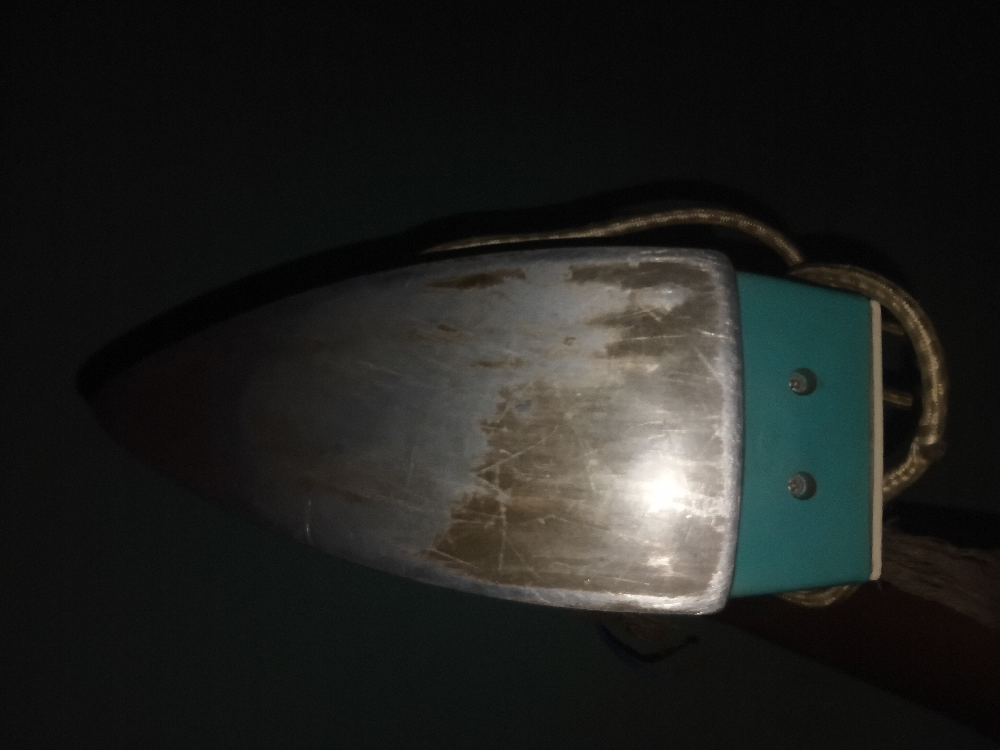 The image shows the top of an old, metal iron with a teal-colored base. The metal soleplate of the iron is scratched and appears to have some discoloration. Two visible screws are on the front of the teal base. A coiled cord, possibly made of braided material, is wrapped around the base and extends out of the frame to the right. The background is dark and out of focus, suggesting a dimly lit environment. The image is a close-up, frontal view of the iron. There is no identifiable location in Jalingo, Nigeria, within the provided image.