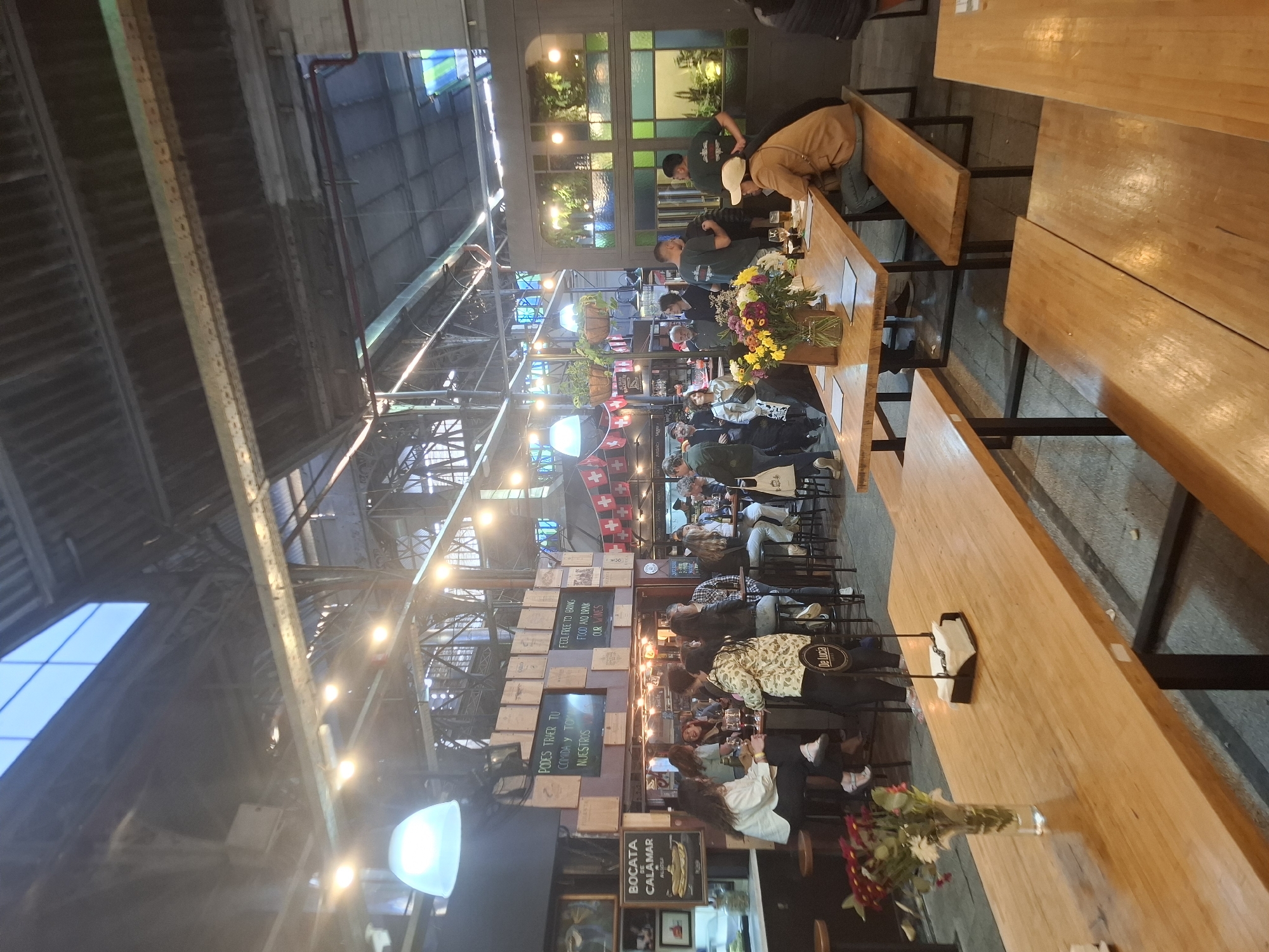 Here is a detailed description of the image:
**Setting:** The image shows the interior of a large, industrial-style building with a high ceiling and exposed metal framework. The space appears to be a bustling marketplace or food hall, filled with people and various food vendors.
**People:** Numerous people are visible throughout the scene. They are dressed in casual clothing and appear to be customers. Some are seated at long wooden tables, eating and drinking. Others are standing, possibly waiting in line or browsing the different vendors. There are also a few individuals who may be vendors or staff, distinguishable by their aprons or uniforms.
**Objects:** Long wooden tables with metal frames dominate the foreground. Some tables have flowers in vases. There are chairs and benches around the tables, providing seating. The vendors have various displays with food and beverages. The walls are decorated with signs, menus, and posters.
**Actions and Interactions:** People are actively eating, drinking, and socializing. Some individuals are engaged in conversation, while others seem to be observing the scene. Vendors appear to be serving customers and preparing food.
**Environment:** The building has a spacious and airy feel. The high ceiling and exposed metal framework create an industrial aesthetic. The lighting is a mix of natural light coming through windows and artificial lights hanging from the ceiling. The overall atmosphere is lively and energetic.
**Location:** Based on the signage that reads "BOCATA CALAMAR", "PODES TRAER TU COMIDA Y TOM NUESTROS", "FEEL FREE TO BRING FOOD AND DRINK OUR WINES", and "De Lucia", it is possible that this is the San Telmo Market (Mercado de San Telmo) in the city of Buenos Aires, Argentina.