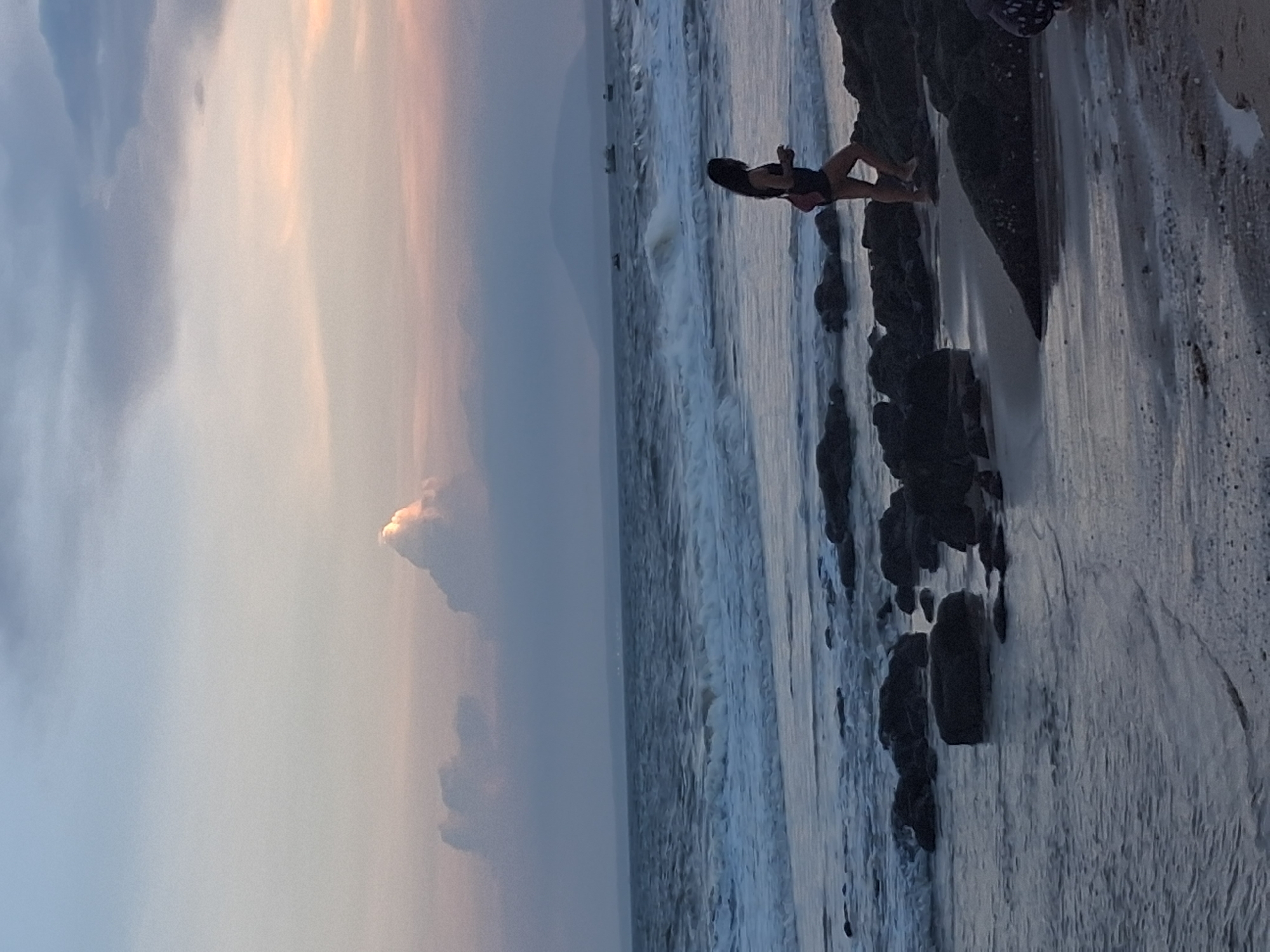 A person stands on a rocky shore with the ocean waves crashing around them. The person is wearing a dark swimsuit with a pink accent. The sky is filled with soft, diffused light, suggesting either sunrise or sunset, with pastel colors blending into the clouds. The ocean is a dynamic mix of white foam and darker water, with waves rolling onto the shore and around the rocks. The rocks are dark and irregularly shaped, with some appearing wet from the tide. The scene captures a moment of solitary connection with nature, perhaps contemplation or enjoyment of the coastal environment. The lighting and atmospheric conditions evoke a sense of tranquility and the natural beauty of a beach at dusk or dawn.