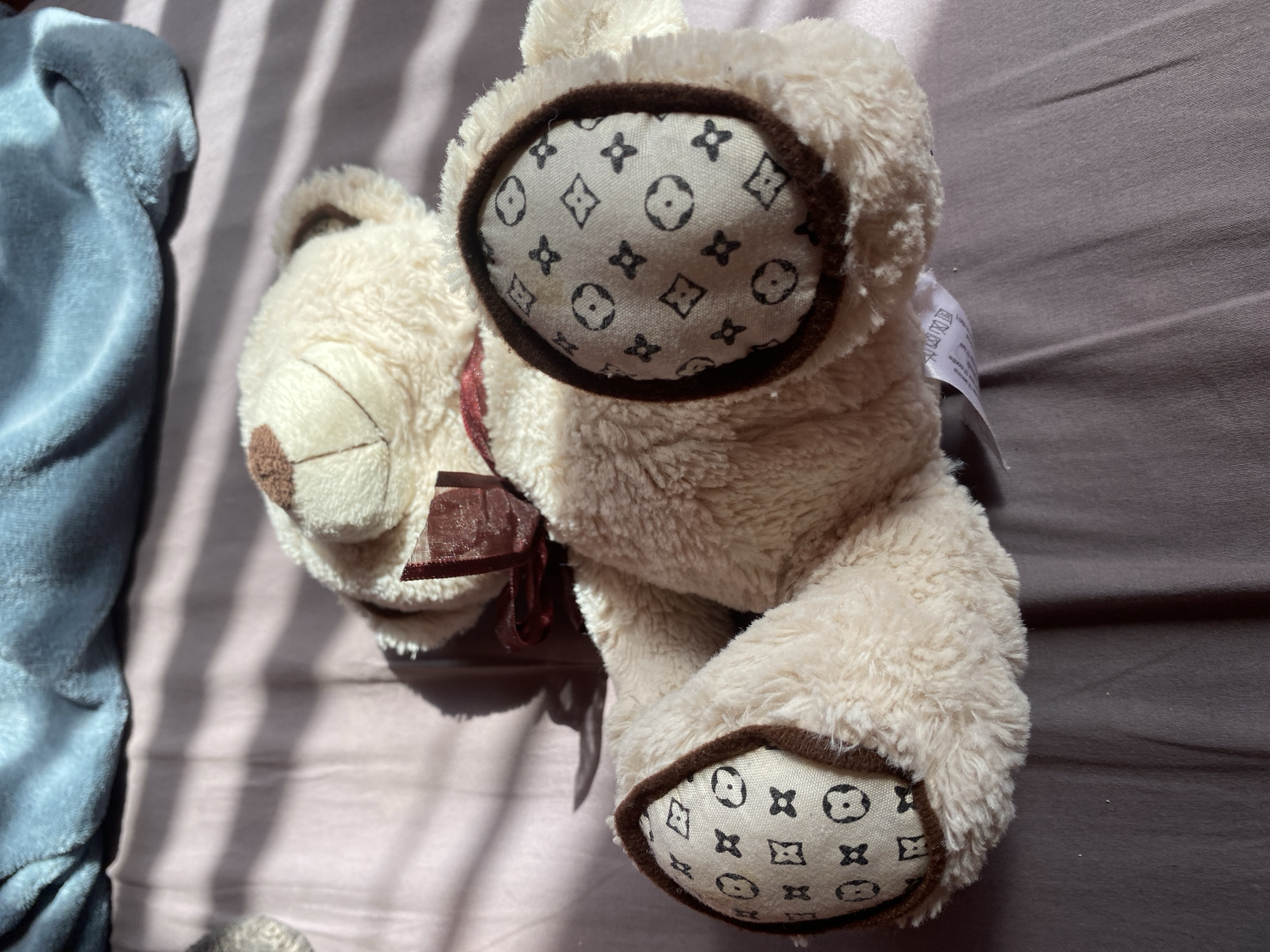 A light-colored, fluffy teddy bear lies on its side on a brownish-grey wrinkled sheet, illuminated by strong, striped shadows suggesting direct sunlight through blinds. A soft, light-blue blanket is partially visible on the left side of the frame.
The teddy bear, cream or beige in color, features a stitched smile and a small brown nose. A dark red or maroon sheer ribbon is tied in a bow around its neck. Its paw pads and the insides of its ears are made of a lighter fabric patterned with repeating symbols resembling the Louis Vuitton monogram: four-pointed stars, circles containing four-petaled flowers, and diamonds enclosing circles. These distinct patterns are framed by a dark brown trim.
A white manufacturer's tag is partially visible near the bear's back, displaying several care symbols including wash at 40 degrees Celsius, do not bleach, tumble dry low, do not iron, and do not dry clean. The setting appears to be indoors, possibly on a bed or a casual resting surface, in Troyan, Bulgaria. No specific activity is taking place beyond the placement of the teddy bear.