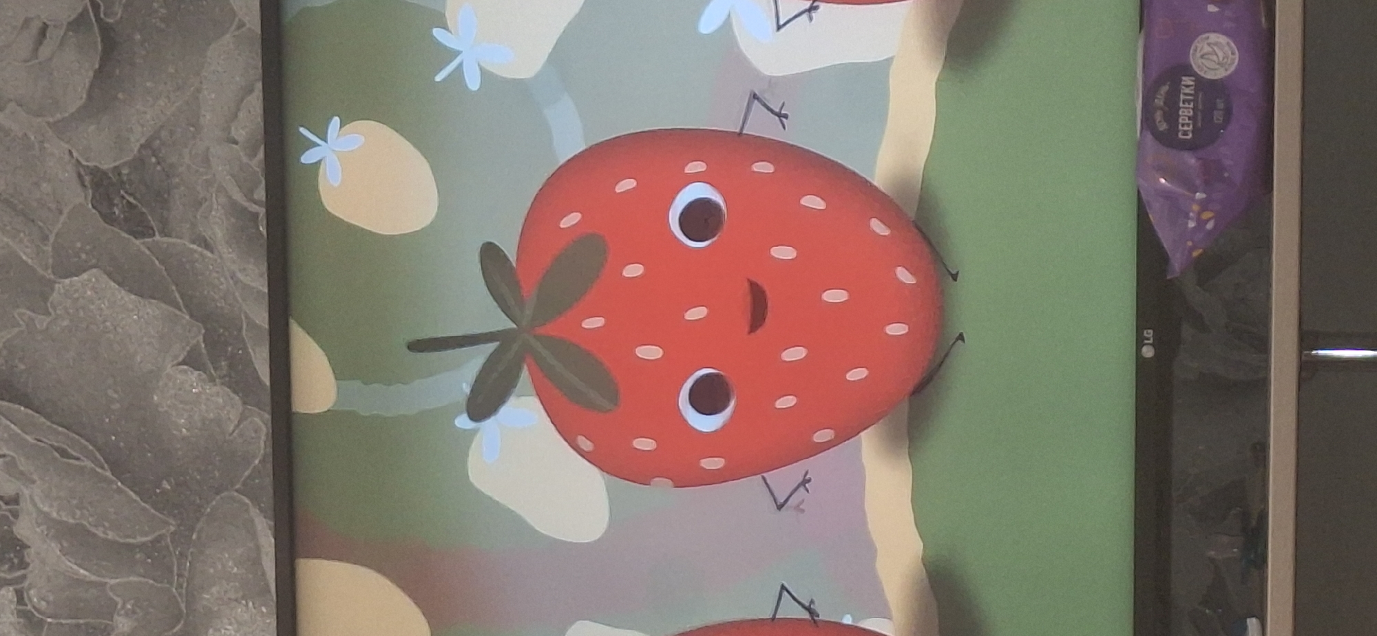 The image shows a cartoon strawberry with eyes, a mouth, and thin limbs. It is positioned in the center of the frame against a green background with abstract yellow shapes and light blue flower-like figures. The strawberry has a stem and leaves on top and appears to be standing on a yellow and green surface. In the lower right corner of the image, a purple package with Cyrillic text, possibly wet wipes, is visible resting on a shelf below the screen. The overall scene suggests a digital display or a picture shown on a screen. There is no indication of a specific city or location within Bila Tserkva, Ukraine.