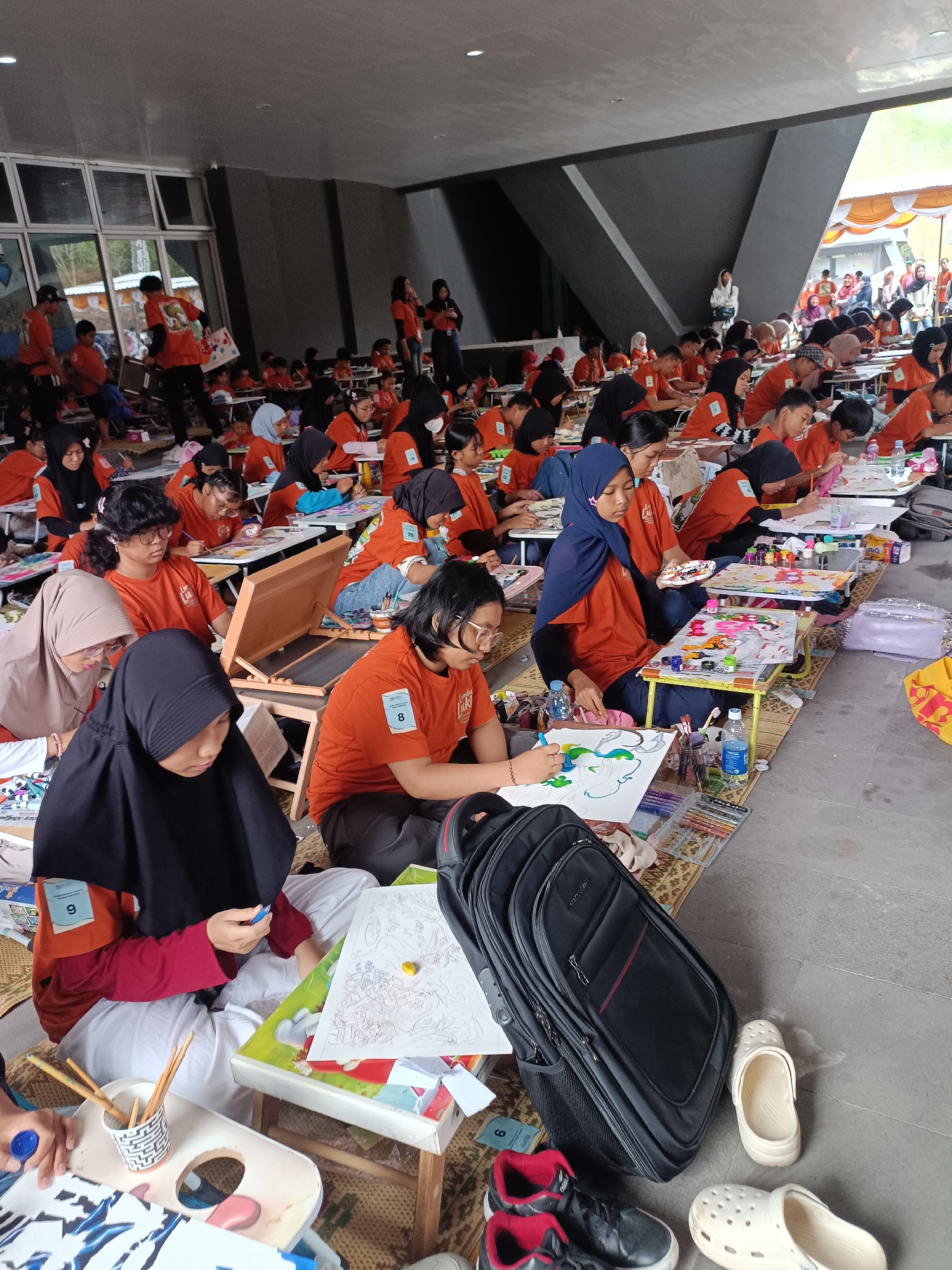 The image depicts an outdoor art competition for children in Hargobinangun, Indonesia. A large group of children, primarily wearing orange t-shirts with the text "Lomba Lukis" (Painting Competition), are seated on the ground, concentrating on their artwork. They are provided with individual low tables for their drawing supplies, including paints, brushes, crayons, and paper. Some children are actively painting or drawing, while others appear to be in contemplation. Various art supplies and personal items like backpacks and shoes are scattered around the participants. The setting suggests a community event, possibly in a park or open space, with a structured arrangement of participants and their workstations. The background shows a building with large windows and a covered area, indicating a semi-shaded environment.
