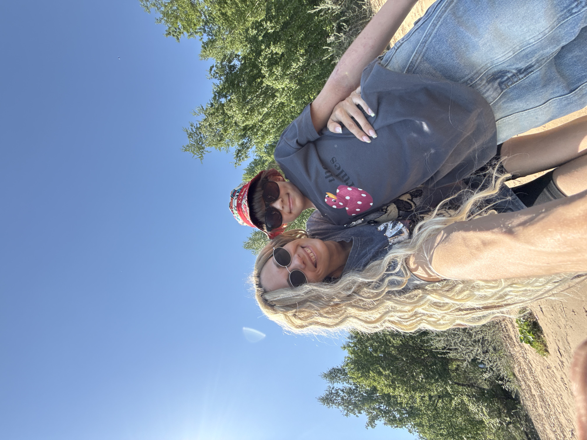 Two individuals are centrally positioned in the frame, appearing to be engaging in a selfie. The foreground features a blonde-haired person with sunglasses, smiling into the camera. Behind them is a younger individual also wearing sunglasses and a patterned bucket hat. Both are wearing t-shirts. The setting includes trees, a clear blue sky, and what appears to be sand or earth near a grassy area. The exact location is not discernible from the given image.