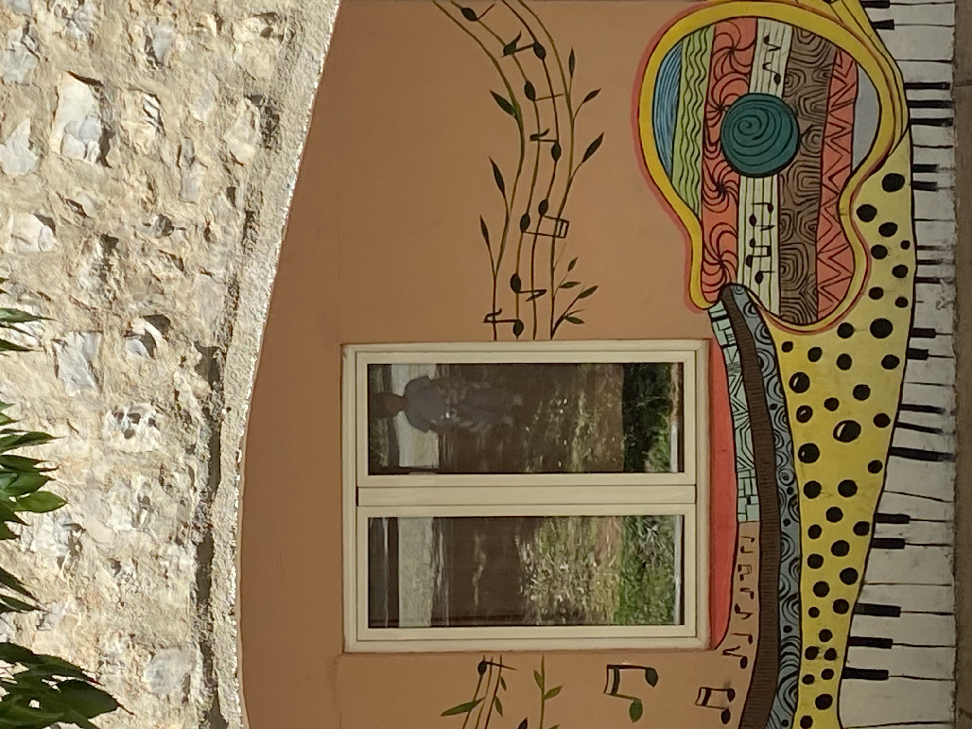 A close-up shot captures a beige wall adorned with a vibrant musical-themed mural. The mural features musical notes on a staff, and a stylized guitar with piano keys on its side. A small, white-framed window reflects an outdoor scene with greenery and a figure, hinting at a passerby. To the left, a textured stone wall adds contrast. This image is likely located in Budva, Montenegro, given the distinct architecture and artistic style often found in the city's public spaces.