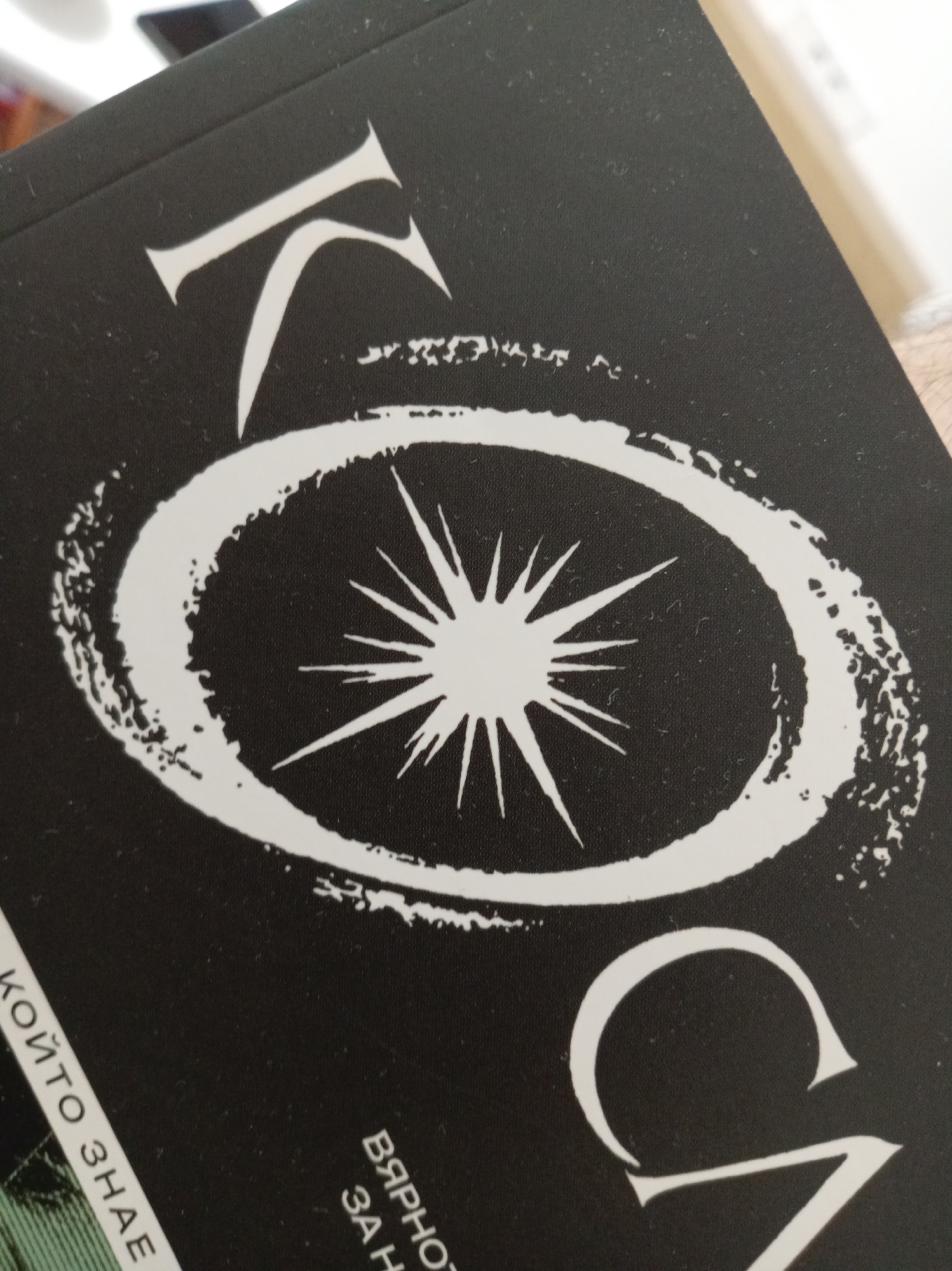 The image is a close-up shot of the cover of a book with a black background and white lettering. The title appears to be in a stylized font, with large, decorative characters. In the center of the cover, there is a circular graphic with jagged edges, enclosing a starburst or sun-like symbol. Some text is visible along the bottom and left edges of the book cover. The text "КОЙТО ЗНАЕ" is partially visible along the bottom left. Towards the bottom center, the text "ВЯРНО" and "ЗА Н" are also visible. The lighting suggests the image was taken indoors with natural light coming from the upper right. The book is tilted slightly, and a person's arm or hand is visible on the right side of the frame, appearing to hold the book. The image quality is moderately sharp, with some dust or debris visible on the book's surface. No specific city or location is identifiable from the image.