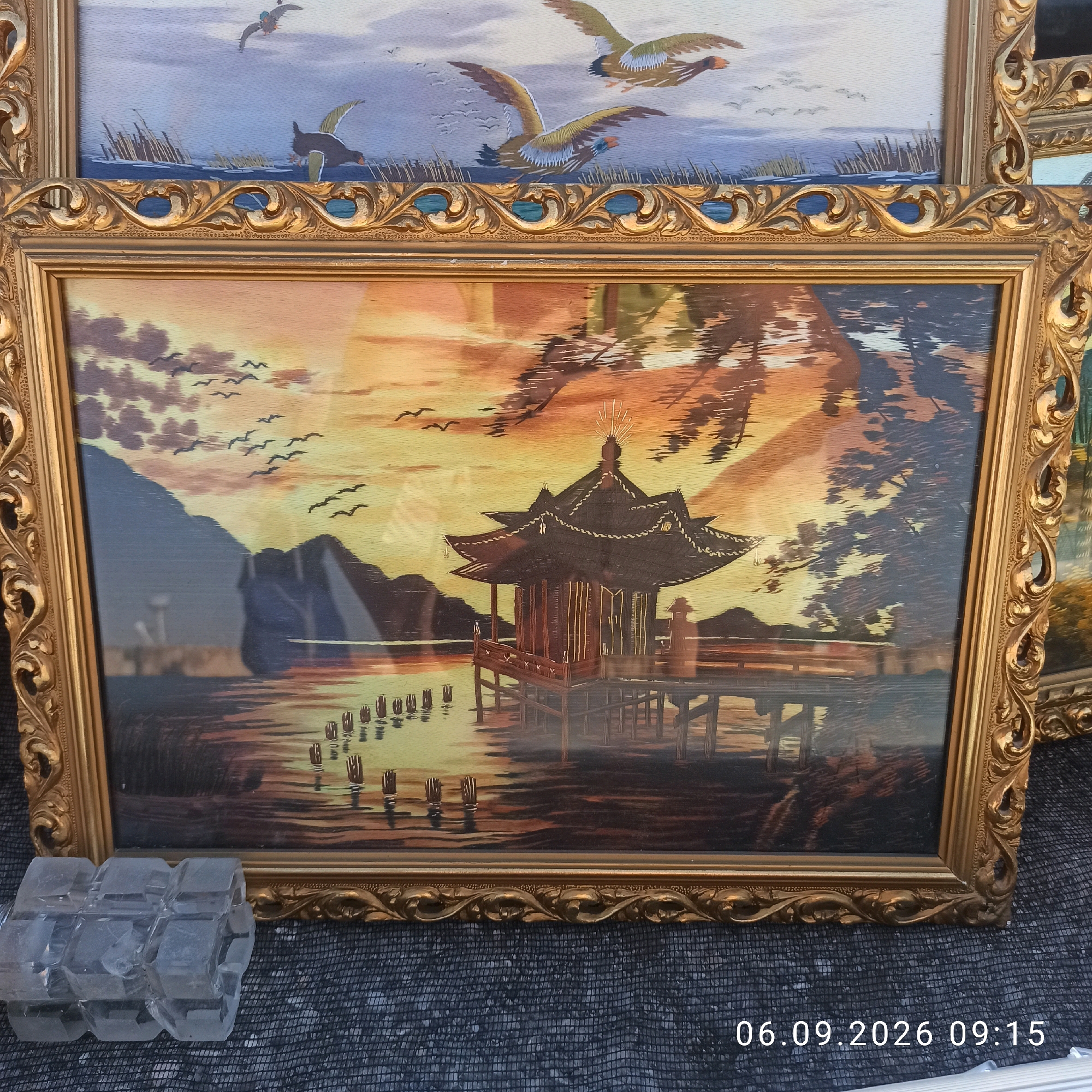 The image displays two framed artworks. The top artwork depicts a scene with waterfowl flying over a body of water with reeds. The bottom artwork is a landscape featuring a traditional Asian-style building on stilts over water, with mountains in the background and birds flying in the sky. Both artworks are framed in ornate gold-colored frames. The bottom artwork appears to be a wood inlay piece. In the foreground, to the left of the bottom frame, is a clear, faceted object that resembles an ice cube tray. The artworks are displayed outdoors, possibly at a market, with a dark, textured surface visible beneath them. A timestamp "06.09.2026 09:15" is visible in the lower right corner of the image. The specific location in Sannicandro di Bari, Italy, is not discernible from the image itself.