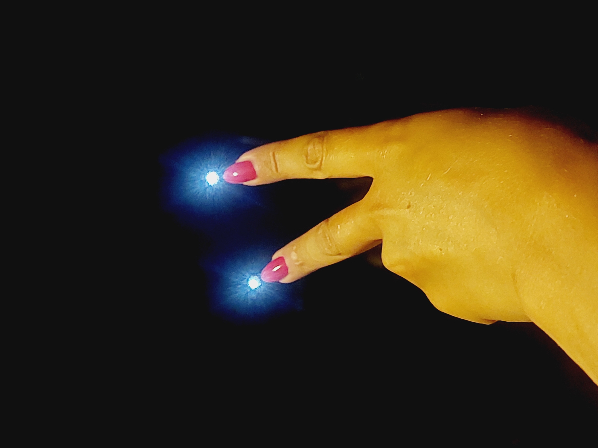 The image shows a hand with bright pink fingernails forming a peace sign against a black background. Two points of bright blue light, possibly lens flares or an artistic effect, are positioned above the fingertips. The lighting on the hand is a warm yellow, suggesting artificial illumination. The location of Sozopol, Bulgaria is not depicted in this image.