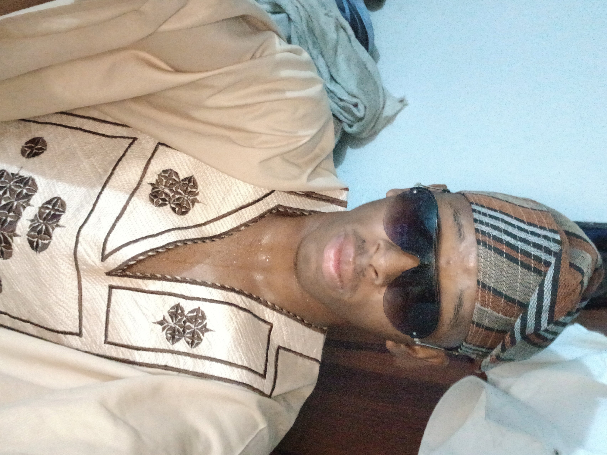 A young Black man, wearing a tan and brown patterned kufi, aviator sunglasses, and a tan dashiki with dark brown embroidery, poses for a selfie. The man's clothing appears to be made of a light, possibly linen-like fabric, and is sweat-dampened around his neck and chest. The background is a plain, light blue wall with some piles of laundry or fabric to the left, and a dark brown surface, possibly a wooden wardrobe, to the right. The image is an indoor portrait. The location is stated to be in the city of Lagos, Nigeria, but this is not visually verifiable from the image alone.