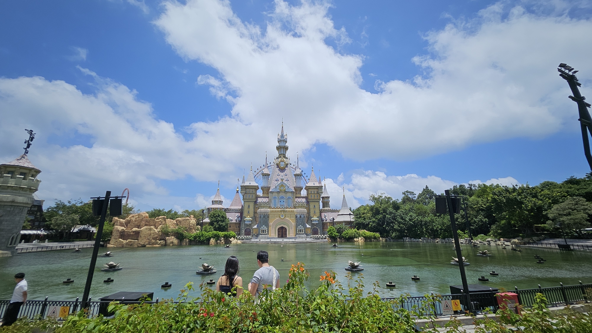 This is a wide-angle, eye-level, outdoor photograph of a fairytale-style castle on a body of water. The castle has a light yellow facade with a pink roof and multiple spires. In front of the castle is a large, greenish body of water with several dark, square objects floating on it, possibly decorative fountains or sculptures. Two people, a woman in a black tank top and a man in a grey t-shirt, are standing in the foreground facing away from the camera, looking at the castle. They are positioned behind a lush green fence with some orange flowers in bloom. To the left of the castle, a tall, grey tower with a pointed roof and decorative elements is visible. The background consists of a dense line of green trees under a bright blue sky with scattered white clouds. The overall scene suggests a theme park or entertainment venue. The location is VinWonders Phu Quoc, Vietnam, an amusement park known for its themed areas, including a fairytale castle that matches the description.