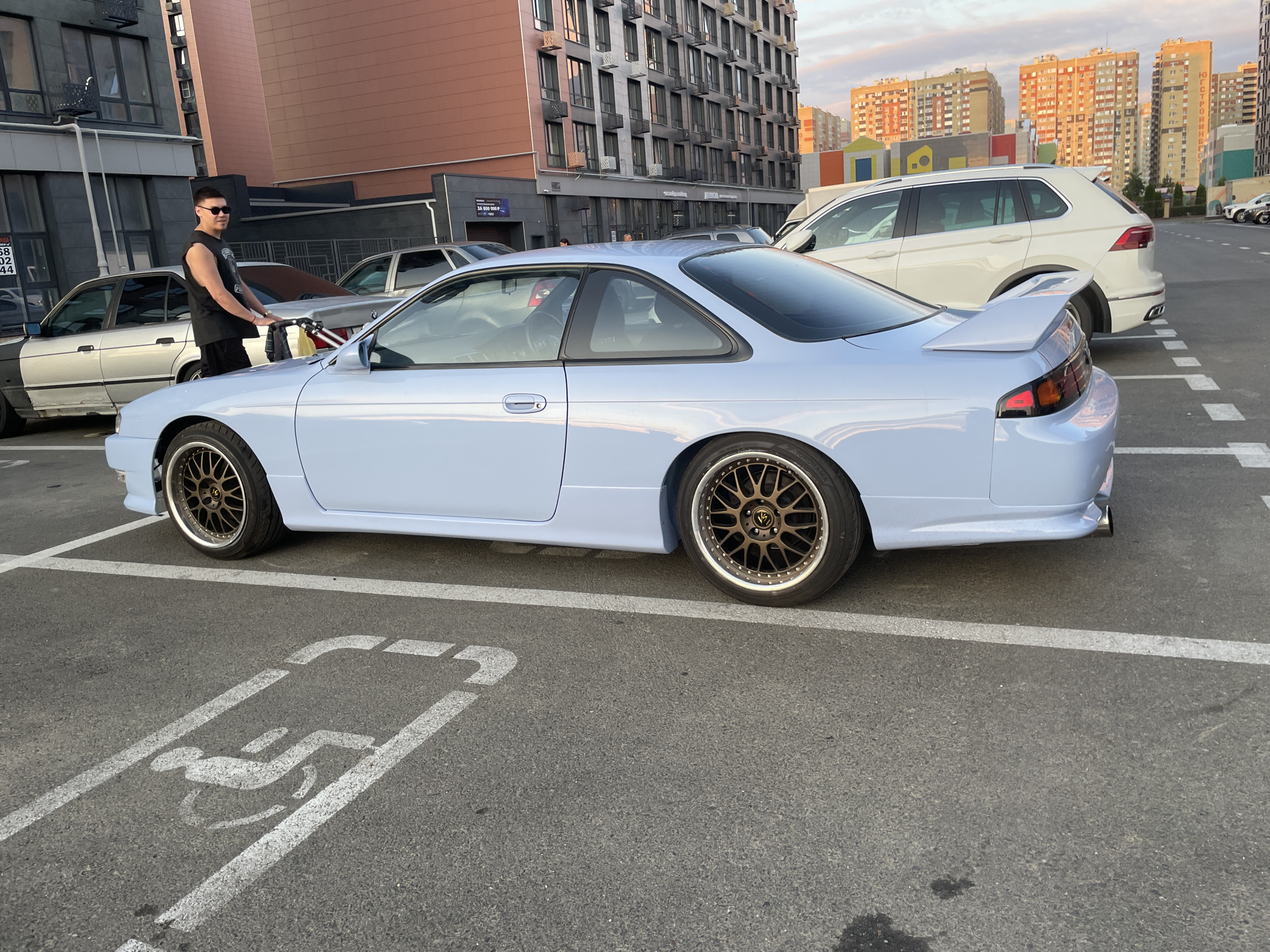 A light blue, two-door coupe is parked in a handicapped parking space. The car has a spoiler and custom wheels. In the background, several apartment buildings are visible, suggesting an urban setting. A person in a black sleeveless shirt and sunglasses stands next to a silver sedan. The pavement in the foreground is marked with white lines, indicating parking stalls. The image appears to have been taken in Stavropol, Russia.