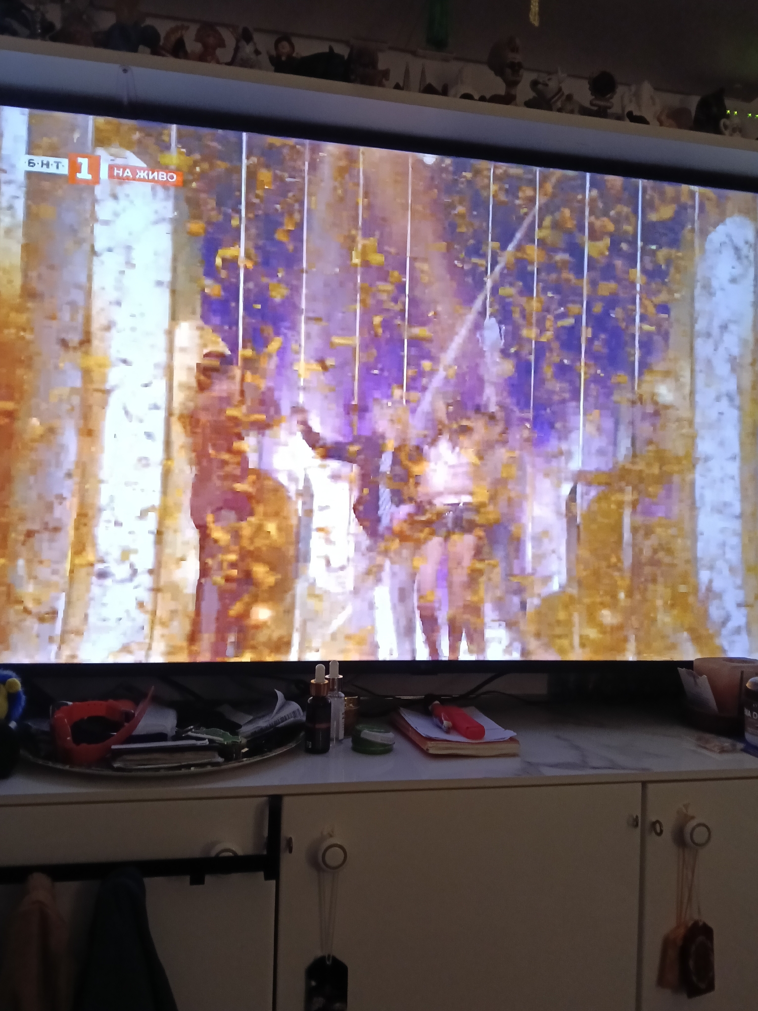 A television displays a celebratory scene, likely a televised event, with confetti falling on a group of people on a stage. The TV is sitting on a white cabinet. In front of the TV on the cabinet are various objects including a red collar, a small bottle of oil, a notebook and a pen. Below the cabinet's surface, items hang from the door handles. The logo "Б.Н.Т 1 НА ЖИВО" is visible in the upper left corner of the TV screen.
The scene appears to be indoors, in a home environment. No details suggest a location in San Bartolomé de Tirajana, Spain.