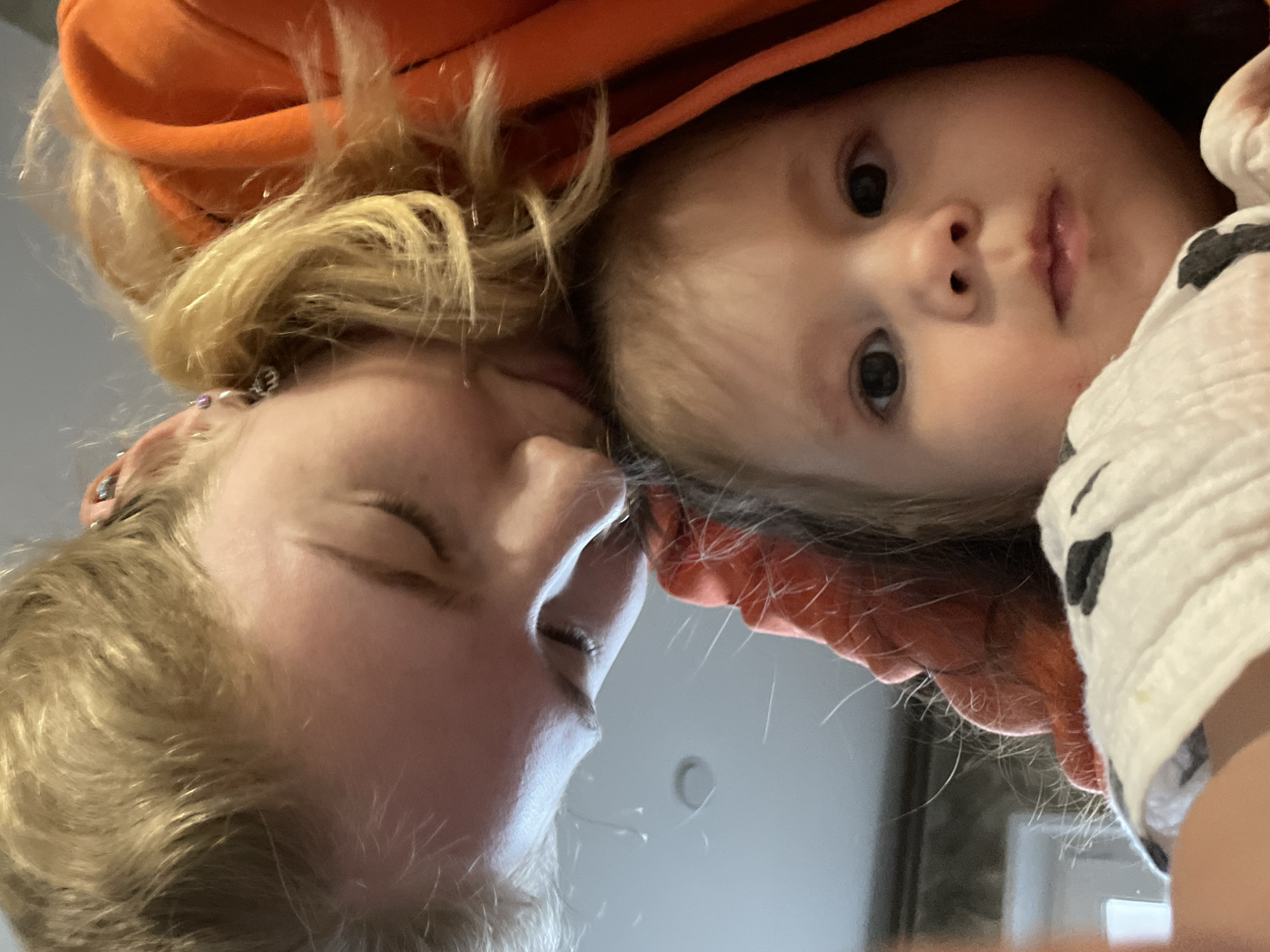 A close-up, low-angle shot features two individuals, likely a woman and a baby, positioned head-to-head. The woman, on the left side of the frame, has blonde hair, her eyes are closed and she appears to be leaning towards the baby. She has visible earrings in her left ear. The baby, on the right side, has lighter hair and is looking directly into the camera with a neutral expression. Both individuals are partially framed by an orange hooded garment. The baby also has a white and black patterned blanket near its head. The background appears to be a plain, light-colored ceiling with a possible light fixture. The image provides no evidence of Izhevsk, Russia.
