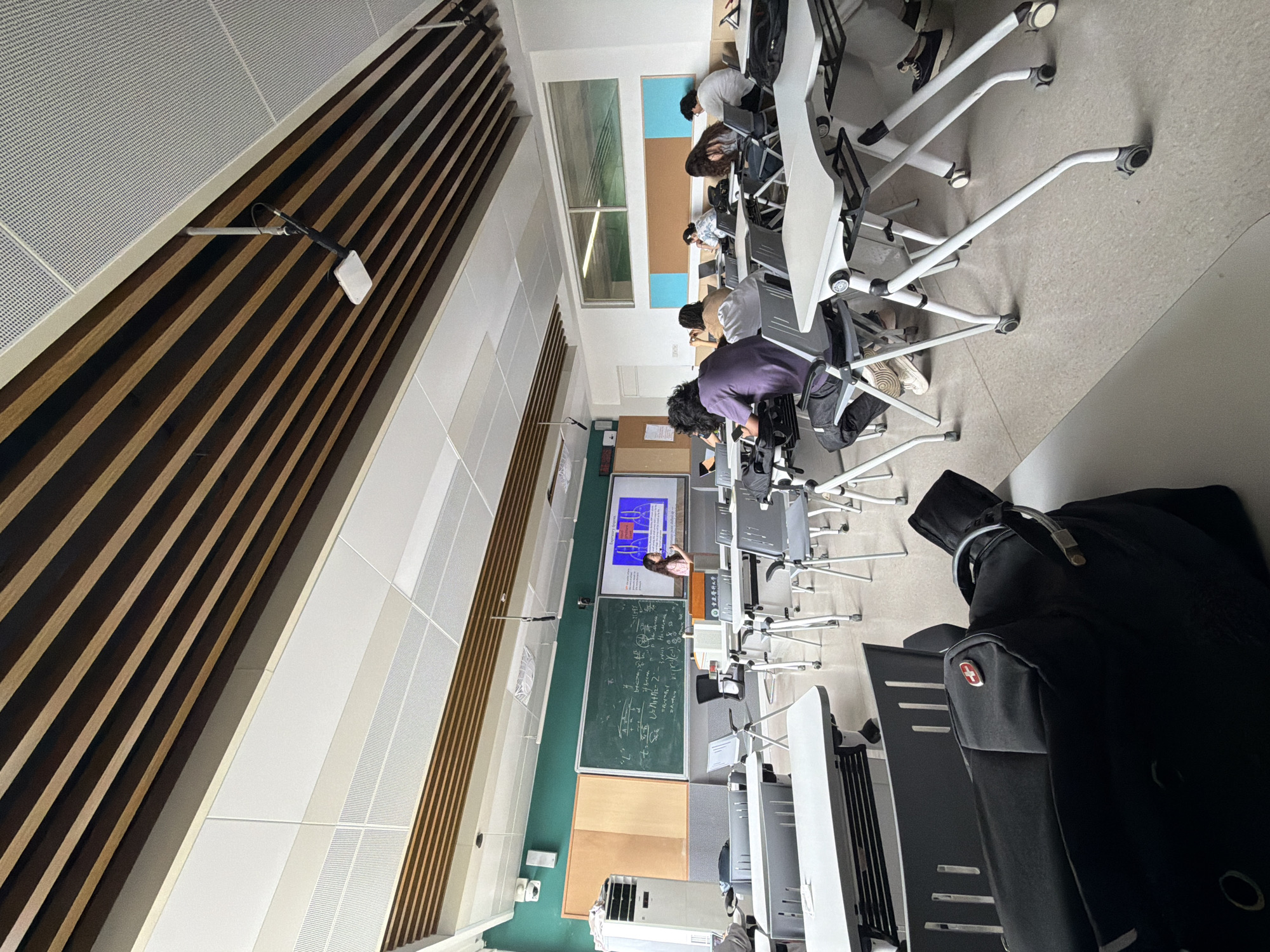 The image shows a classroom in Chongqing High-tech Zone, China. Students sit at desks with built-in chairs facing the front of the room. A blackboard with equations and a screen displaying diagrams are at the front. A black backpack sits on a desk in the foreground. The room has distinct wall and ceiling panels, with a mix of wood and white materials. Sunlight streams through a window on the left.