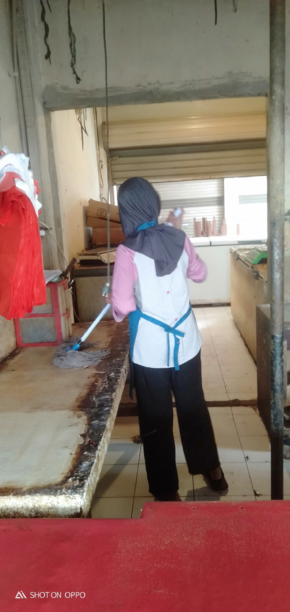 An individual wearing a dark grey hijab, a pink and white shirt, a blue apron, and black pants is mopping a dirty counter with a wet mop. The individual is standing on a tiled floor in what appears to be a commercial kitchen or food preparation area. To the left of the counter, there are stacks of red and white fabric. In the background, there are refrigerators and stacks of boxes. The scene is illuminated by artificial lighting. The overall impression is of a cleaning in progress in a functional, potentially unglamorous, environment.