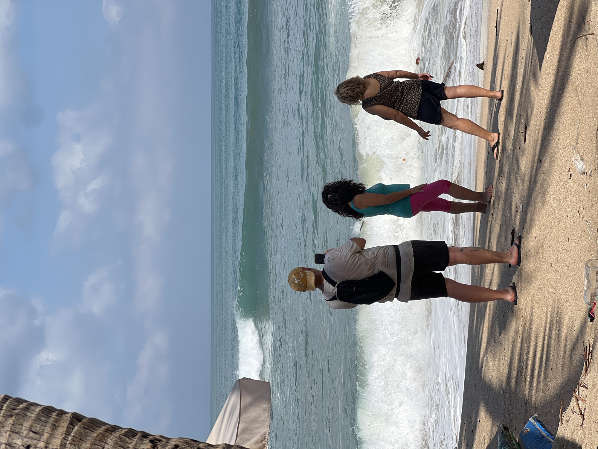 The image depicts a group of three people standing on a sandy beach facing the ocean. A man in the foreground on the left is wearing a light-colored t-shirt, black shorts, a yellow cap, and a black backpack. He is holding a phone and appears to be taking a picture or recording. To his right, two women stand closer to the water. The woman on the left has dark, curly hair and is wearing a teal tank top and pink capri leggings. The woman on the right has blonde, curly hair and is wearing a patterned sleeveless top and dark shorts. They are all looking out at the ocean, where waves are breaking. The water is a turquoise blue, and the sky is partly cloudy. In the background, a large tan umbrella or canopy is partially visible on the left side of the frame. The beach itself has scattered debris, including a plastic bottle and some branches. The image captures a moment of leisure or observation at the seaside, possibly in Kuta, Indonesia, a popular tourist destination known for its beaches and surf.