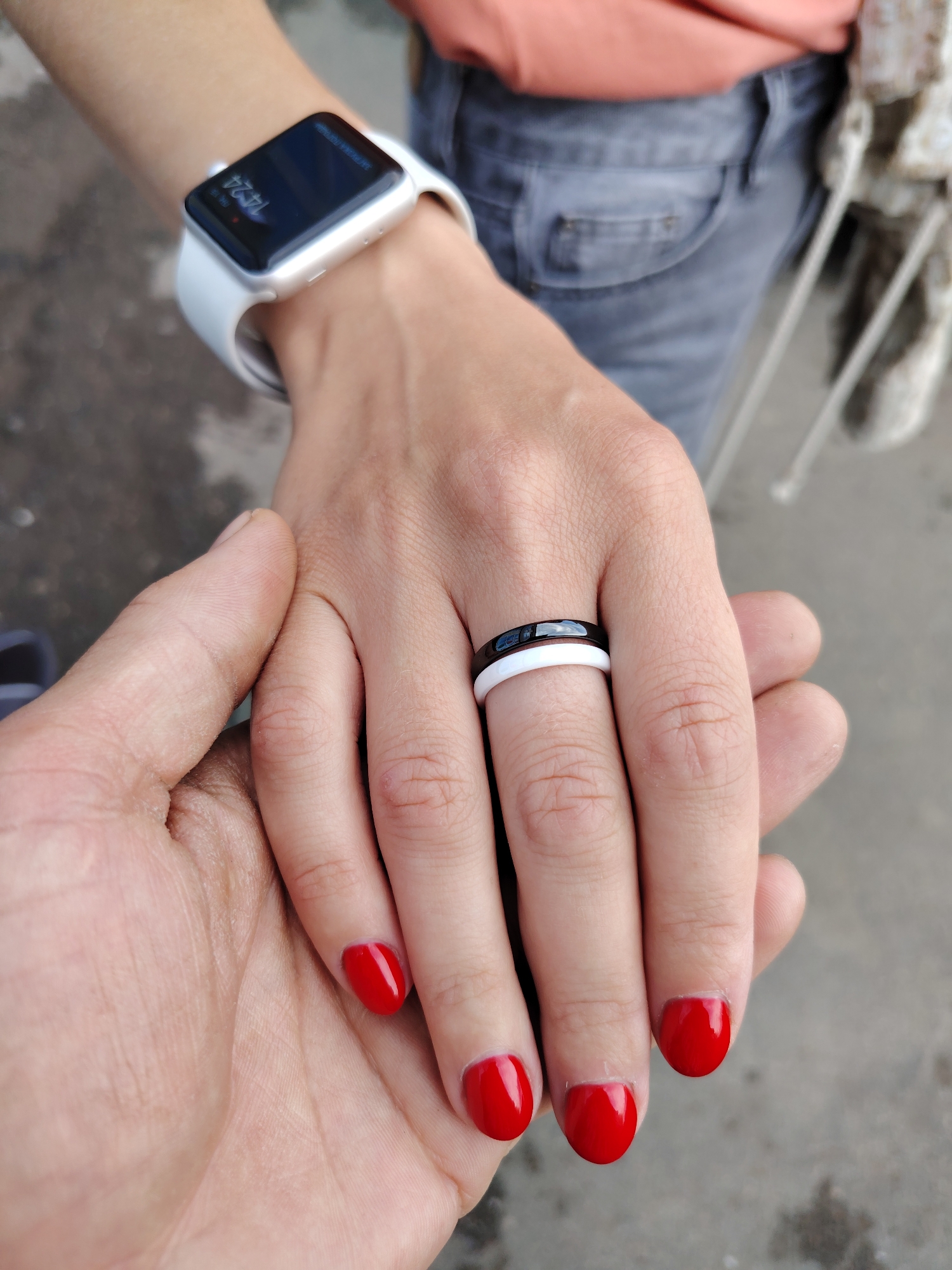 The image is a close-up of two hands intertwined. The hand on the right belongs to a person wearing a white smartwatch and two rings on their finger: a thin black ring on top and a thin white ring underneath. This hand has bright red nail polish. The hand on the left is gently holding the hand of the person on the right. The background is blurred, showing a grey, textured surface, possibly pavement, and hints of clothing that appear to be denim jeans and an orange shirt. There is no discernible location within the city of Chaikovsky, Russia, visible in the image.