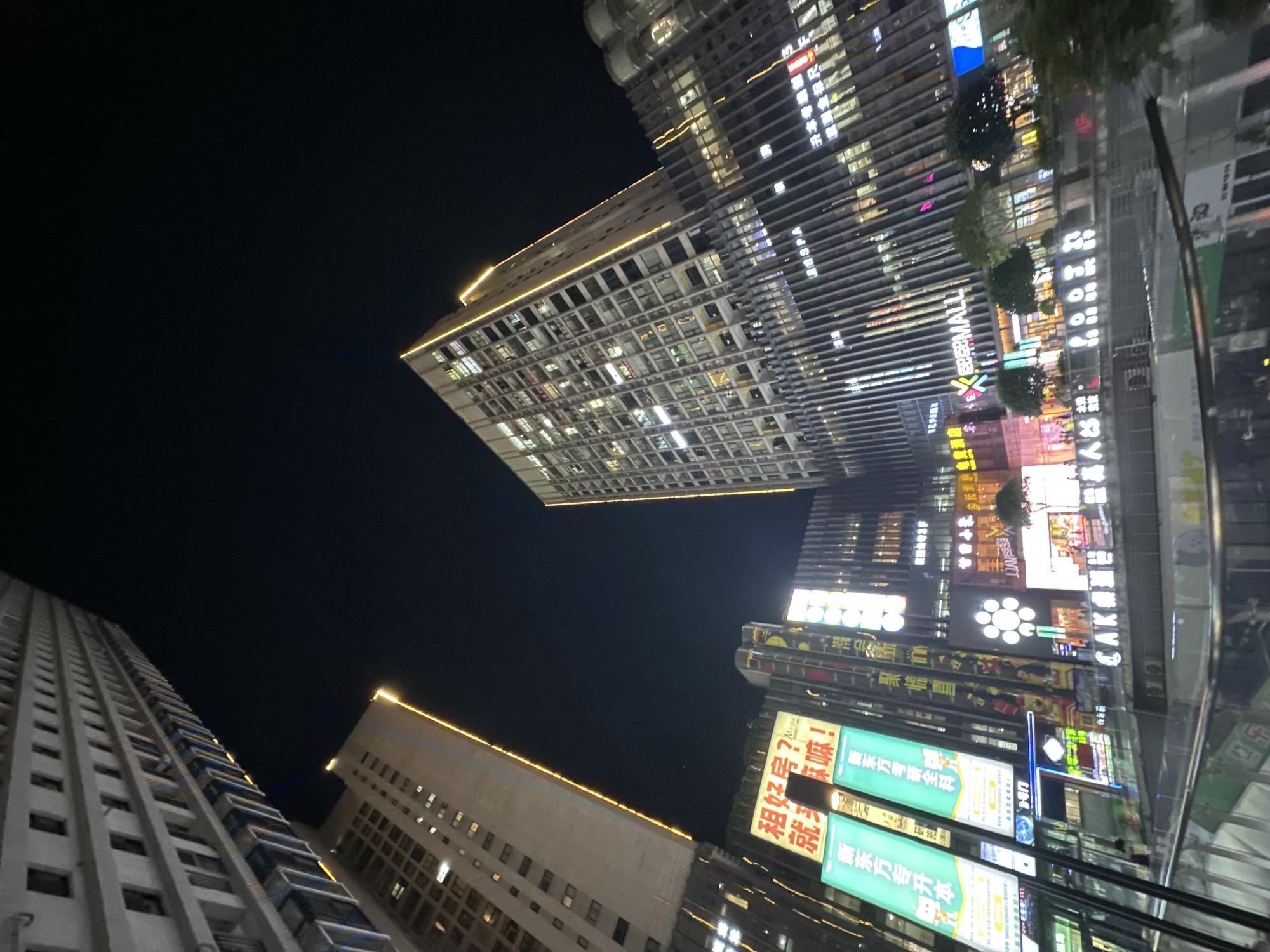 The image depicts a nighttime cityscape in the Chongqing High-tech Zone, China. Several tall buildings are visible, illuminated by internal lights and external signage. Advertisements and commercial names in Chinese are prominent on some of the buildings. The sky is dark, suggesting it is night.