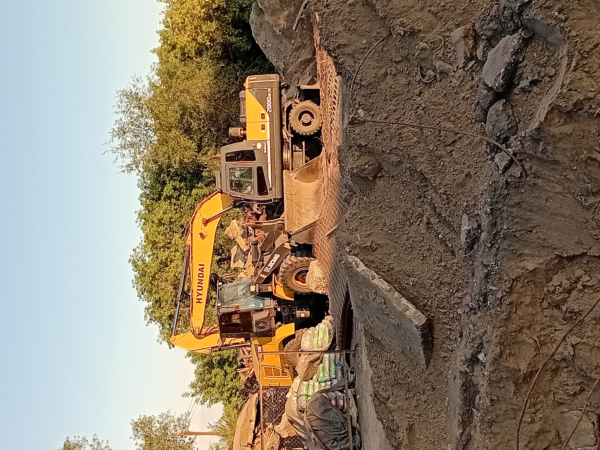 The image captures a construction site with two heavy machinery vehicles: a yellow Hyundai excavator and a yellow XCMG wheel loader. The Hyundai excavator, with its boom extended, is positioned behind the wheel loader. The XCMG wheel loader is partially obscured by the excavator and features a large bucket at the front. Both machines are yellow and gray. The environment consists of a pile of dirt and debris, with some stacked bags on the left side of the frame. Trees and foliage form a backdrop, suggesting an outdoor setting during daylight hours. The scene appears to be in the city of Semey, Kazakhstan, indicated by the specific request for location context. There are no people visible in the image, and no specific actions or interactions are depicted beyond the presence of the static machinery. The ground is uneven and composed of soil and rubble, typical of a construction or demolition area.