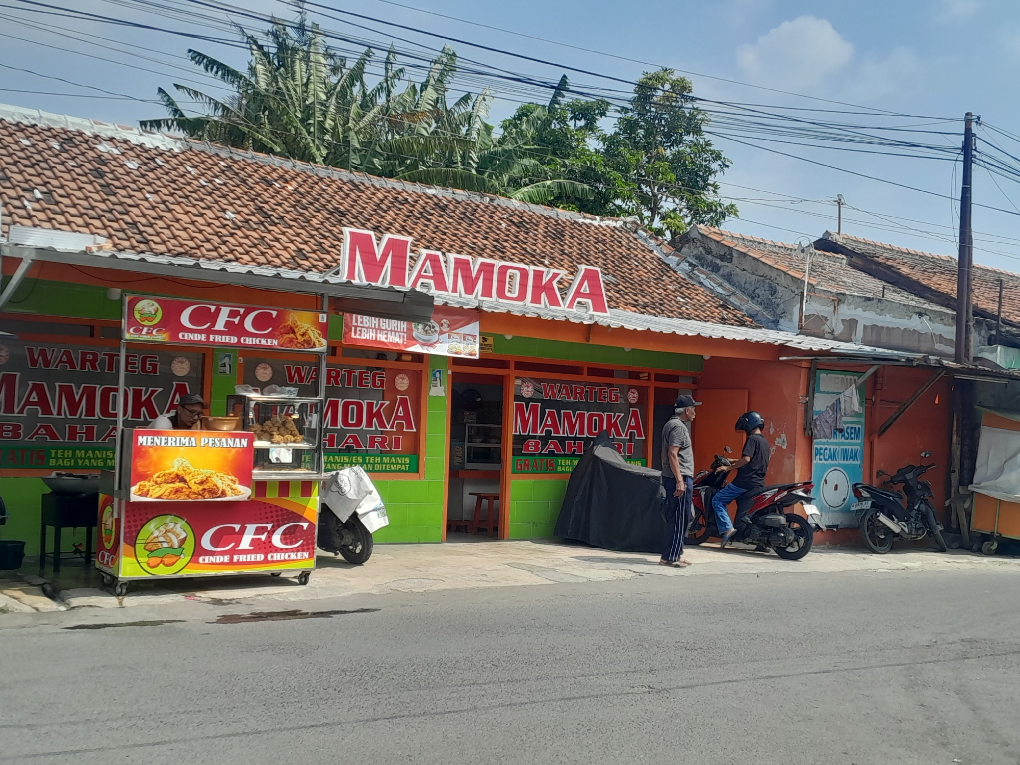 A street-level, outdoor, medium shot captures a commercial establishment in Tegal, Indonesia, on a sunny day. The primary focus is on a food stall with bright signage featuring the name "MAMOKA" in large red letters on the storefront and a cart branded "CFC CINDE FRIED CHICKEN." The stall appears to be selling fried chicken, as indicated by the prominent image on the cart.
Several individuals are visible. One man, wearing a grey t-shirt and dark pants, stands near the entrance, seemingly conversing with another person. Another individual, wearing a black t-shirt and blue jeans, is seated on a red motorcycle. Two other motorcycles are parked nearby, one of which is covered with a black tarp.
The environment includes a paved road in the foreground, with a concrete sidewalk in front of the establishment. The building itself has a terracotta-tiled roof and orange-painted walls. Electrical wires crisscross the sky above. Tropical trees, including banana trees, are visible behind the building, suggesting a warm climate. The scene depicts a typical street-side business in a local Indonesian setting.