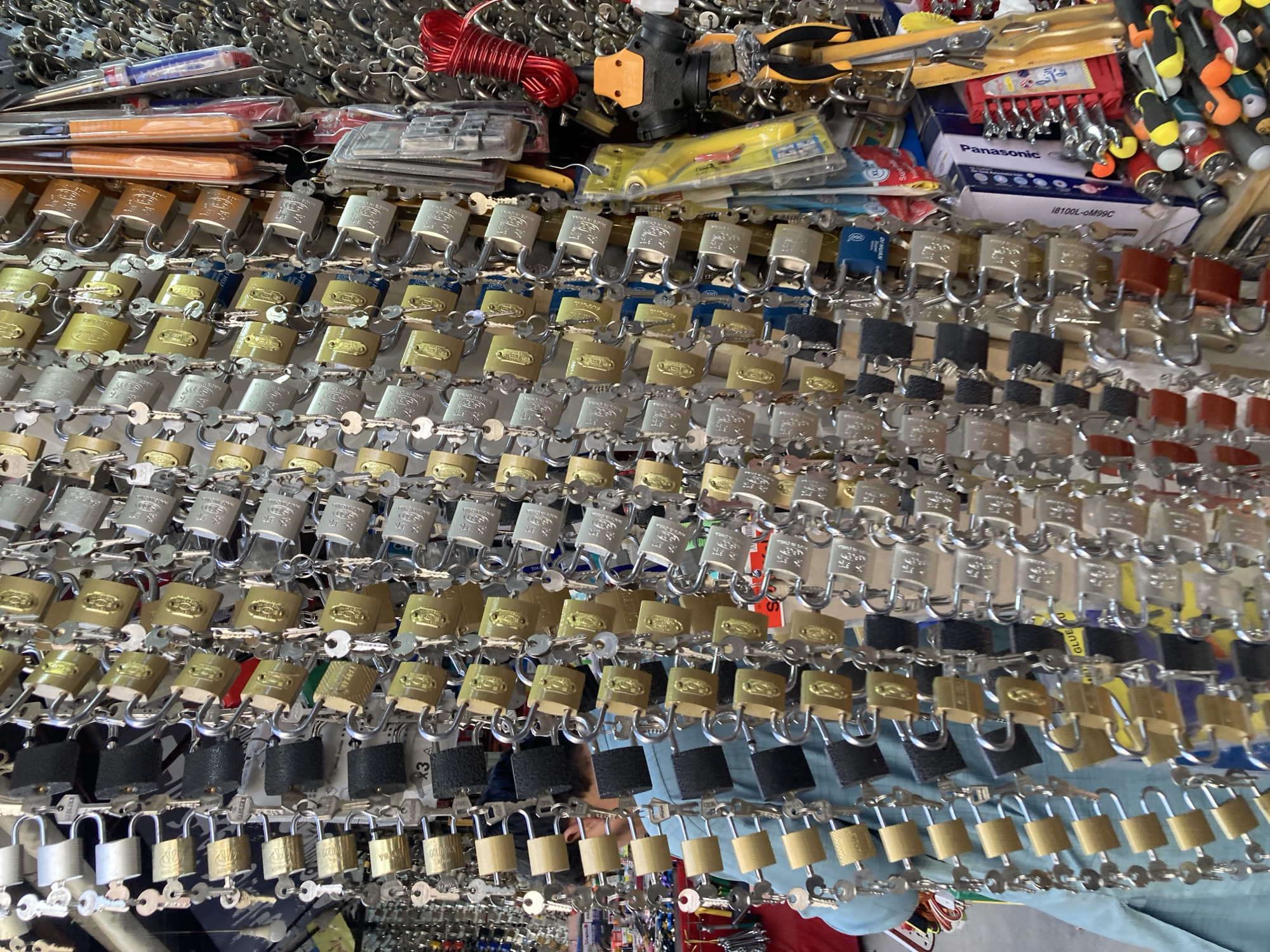 This media file captures a densely packed display of various hardware items, primarily padlocks, in what appears to be a market stall or shop in Quetta City Tehsil, Pakistan.
The foreground is dominated by numerous rows of padlocks of different sizes, colors, and materials. These include golden, silver-toned, brass-colored, black, red, and blue padlocks. Each padlock typically has its corresponding key attached, suggesting they are sold as sets. Many padlocks bear visible text such as "MADE IN CHINA" and brand names like "GOLD" and "DI HUA." Some also have numbers like "3," "84," "X," "63," "200," and "000" or letters like "XL." One padlock also has "JAPAN" inscribed.
Behind and above the padlock display, a variety of other tools and hardware are visible. These include bundled screwdrivers, a red coil of wire, pliers, a black pipe fitting, and various small items in clear plastic packaging. Packaging for "Panasonic" brand items, specifically a box labeled "18100L-OM99C," is also visible. Other text fragments like "GLUE U" and "Su" are present on packaging or labels.
The items are arranged in a retail fashion, with padlocks neatly organized on hooks or chains, and other items piled behind them. The bright lighting suggests either a well-lit indoor space or an outdoor stall during daytime. A faint reflection of a person wearing a blue shirt is visible among the lower padlocks, indicating an active shopping environment. The overall impression is one of a busy, functional hardware vendor.