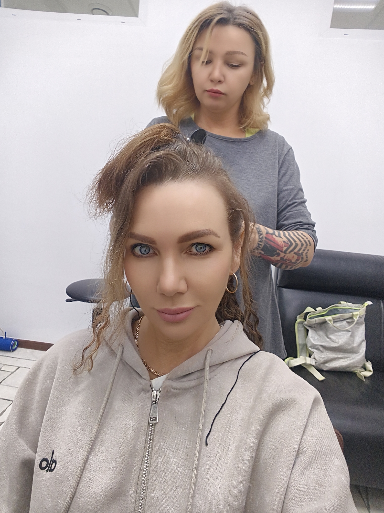 In a brightly lit indoor setting, likely a salon or studio with white walls and a tiled floor, two women are present. In the foreground, a woman with light skin, striking blue eyes, and long, voluminous brown hair (partially styled upwards) is looking directly at the camera with a neutral expression. She wears a light gray or tan zippered hoodie, a gold chain necklace, and gold hoop earrings. On the left chest of her hoodie, the stylized lowercase text "alo" is visible.
Behind her, a second woman, possibly a hairstylist, is attending to her hair. This woman has blonde hair and wears a plain gray long-sleeved shirt. She appears focused downwards, and a prominent, detailed tattoo, featuring intricate patterns and possibly some symbols, covers her right forearm. Sunglasses are tucked into her shirt collar.
The foreground woman is seated in what appears to be a black upholstered chair. To the right, another black chair holds a light gray fabric bag with lime green accents. The overall atmosphere suggests a professional grooming session.