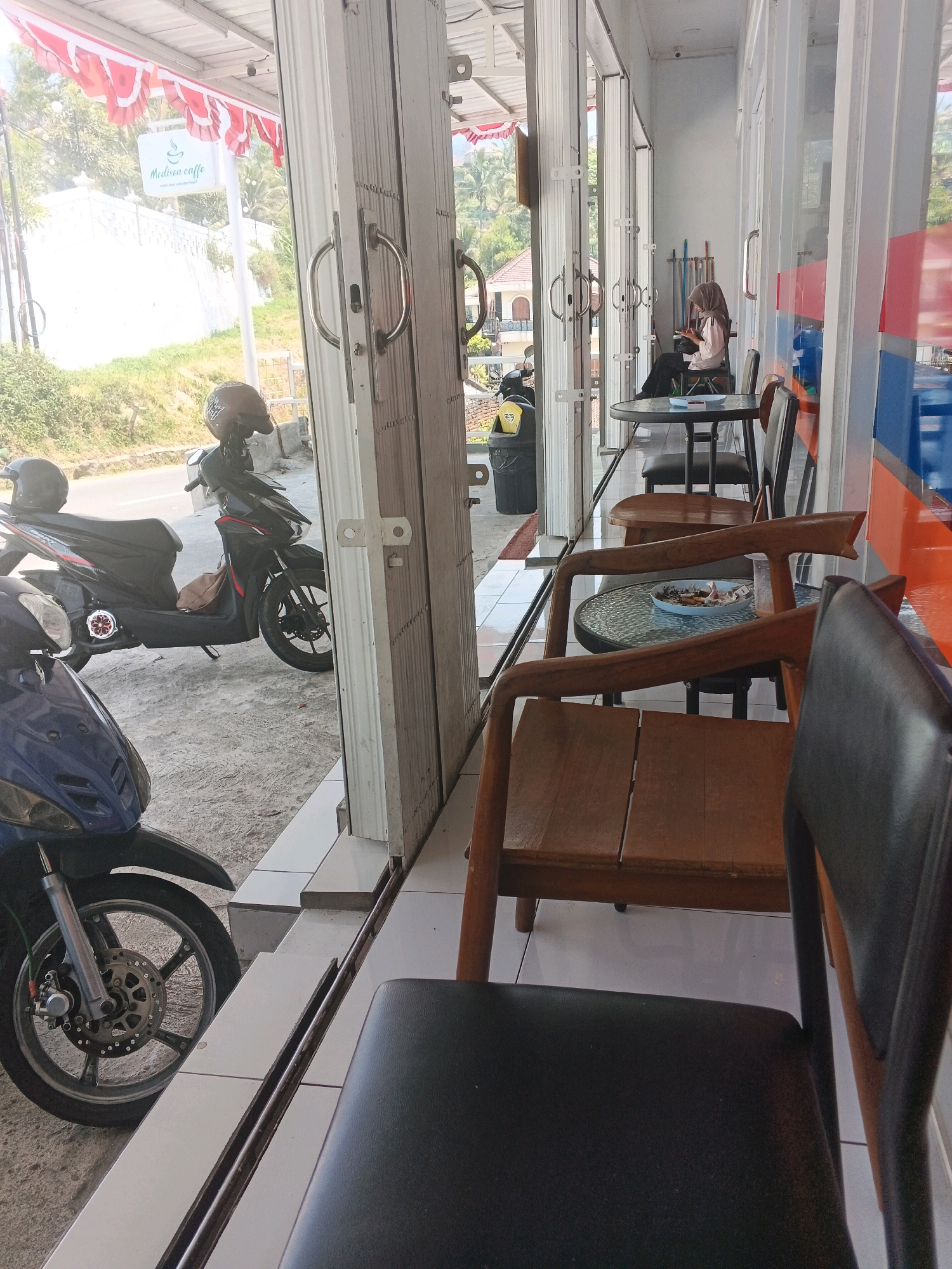 This image depicts the exterior of a cafe named "Madison caffe" in Pakopen, Indonesia. In the foreground, a blue motorcycle is partially visible, with its front wheel and fender in focus. Behind it, a black motorcycle with two helmets and a brown bag on the seat is parked. The cafe features large glass doors that slide open, revealing the interior. A woman wearing a hijab and a light-colored top is seated at a small round table, looking at her phone. There are several chairs and tables outside the cafe. Red and white pennants are strung above the entrance. The cafe is situated along a road with a white wall and lush greenery in the background.