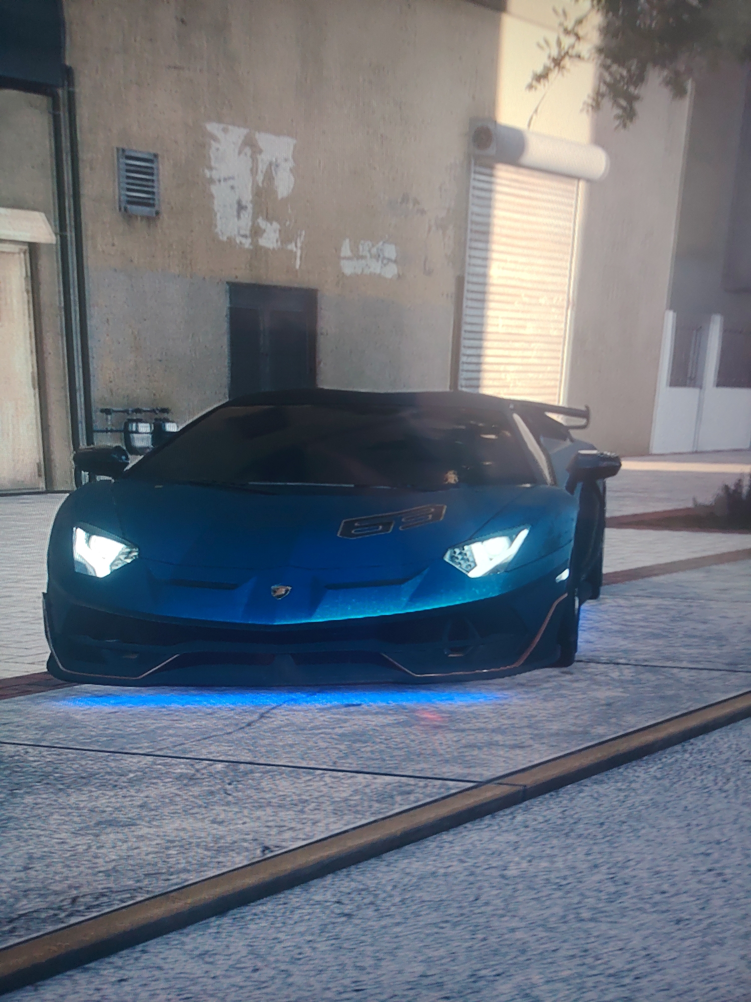 A blue Lamborghini is parked on a concrete surface in front of a beige building with a roll-up garage door. The car has its headlights on, and a blue glow emanates from underneath the front of the vehicle. The background features peeling paint on the building and a small ventilation grate. The location is in an urban area within the city of Unknown, Unknown.