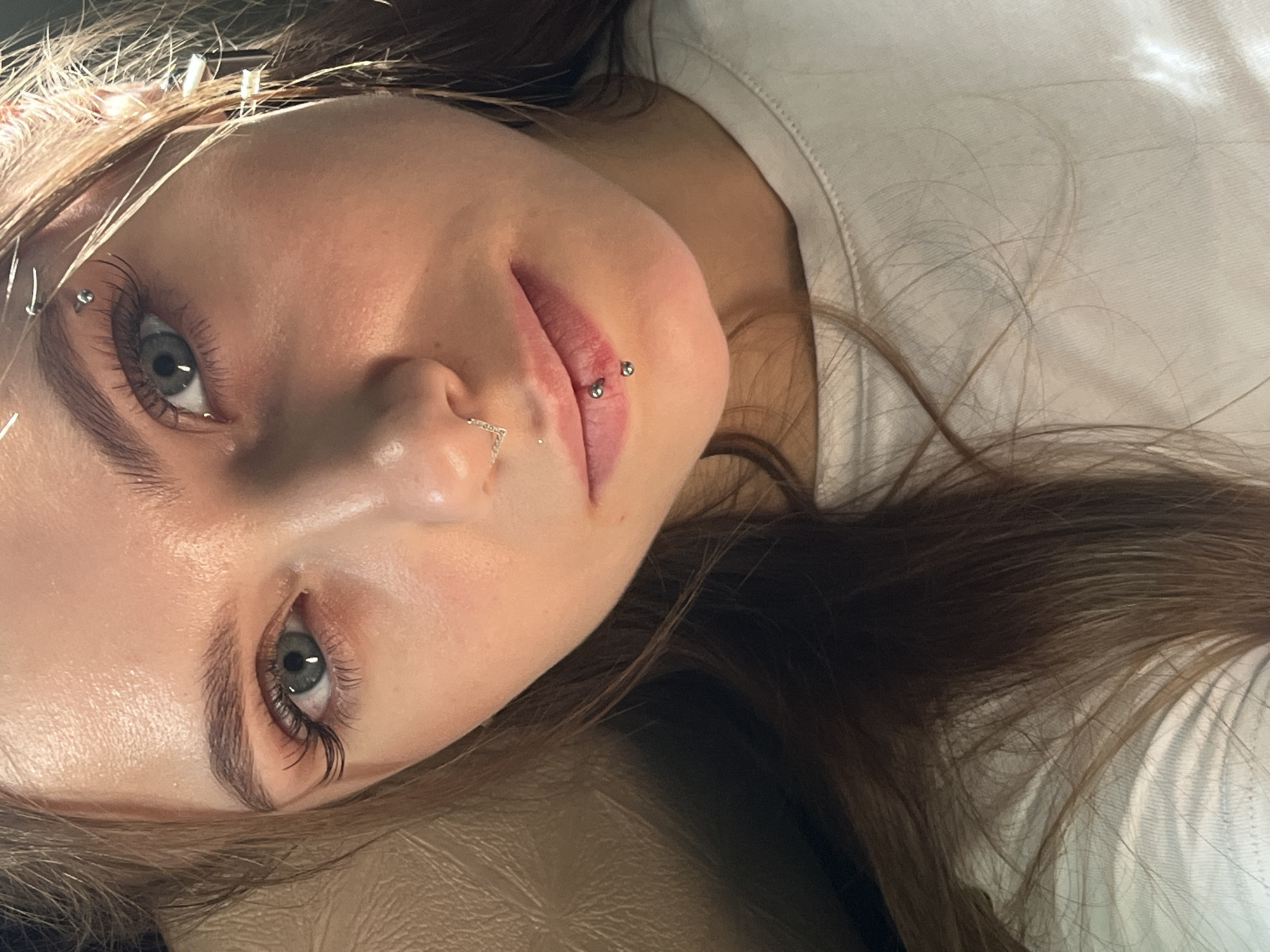 A close-up, slightly angled headshot of a young Caucasian woman. She has light skin, light brown hair, and gray eyes. She has multiple facial piercings: one above the left eyebrow, a ring in the left nostril, and a labret piercing on the left side of her lower lip. She is wearing a white top. The background is a tan color. The location is unspecified.