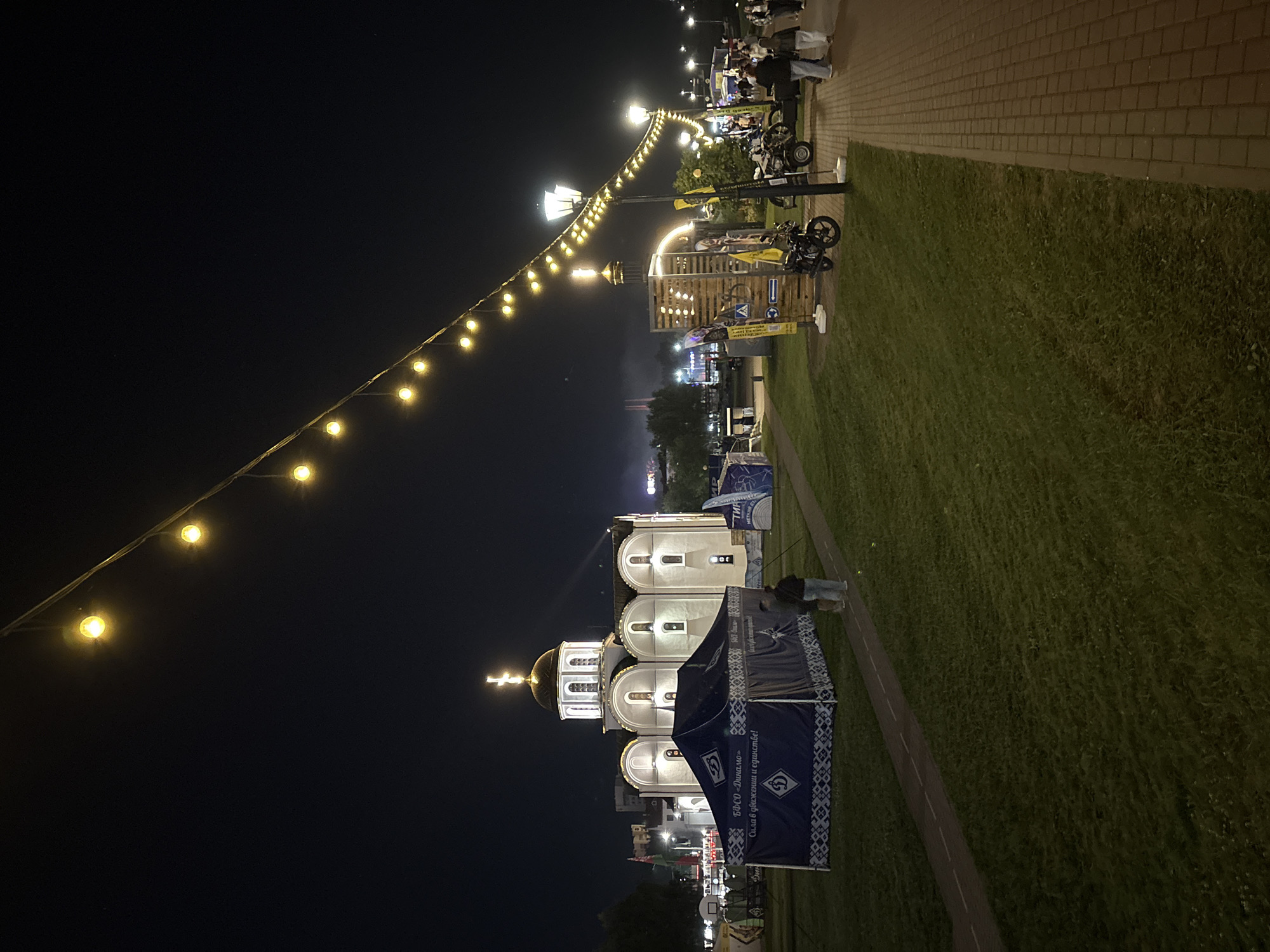 The image depicts an outdoor scene at night in Vitsebsk, Belarus. In the foreground, a brick-paved walkway runs alongside a grassy area. String lights with yellow bulbs are strung diagonally across the dark sky.
To the left, a white church with a golden dome and cross is illuminated. In front of the church, a large blue tent with white patterned trim is set up. Two individuals are standing near the tent, facing away from the camera.
Further into the scene, there are more tents, wooden structures, street lamps, and several motorcycles parked. People are visible walking along the walkway and in the background. In the far background, there are buildings and lights suggesting a city environment.