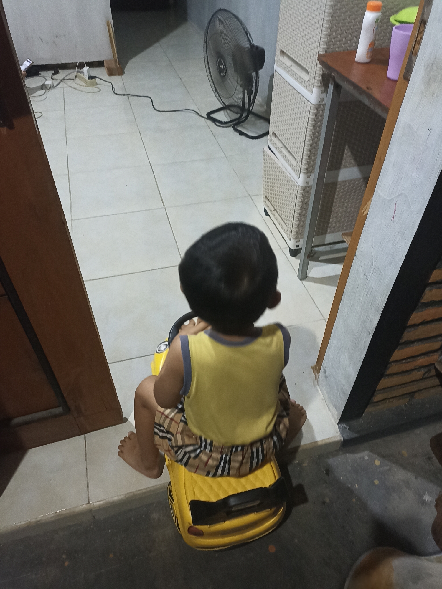 A child sits on a yellow toy car in a room in Mojopanggung, Indonesia. The child is wearing a yellow tank top and plaid shorts. The room has tiled floors and a fan in the background. A white cabinet and a wooden table are also visible.