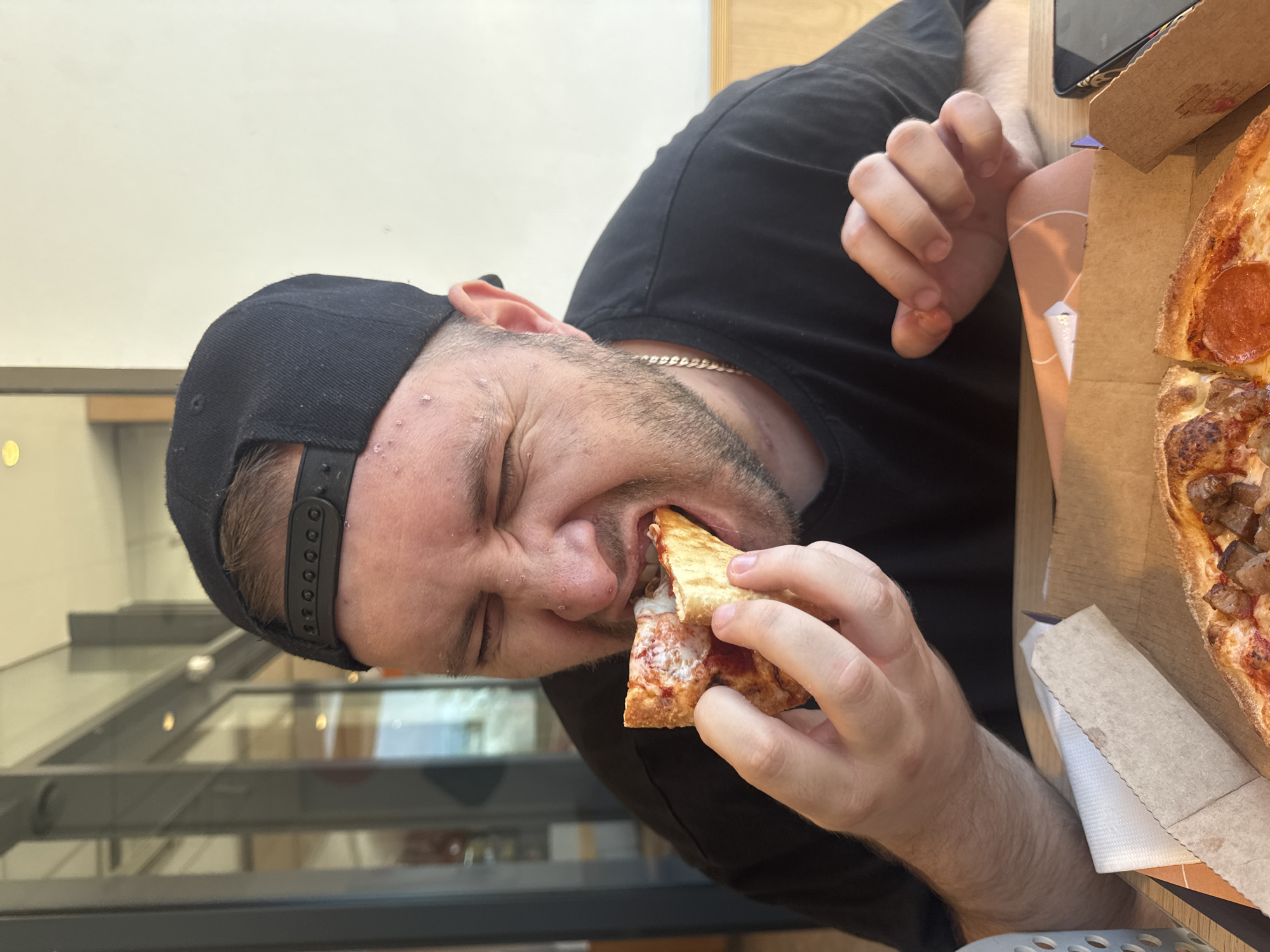 A man wearing a black baseball cap and a black t-shirt is eating a slice of pizza. He is holding the pizza slice with his left hand and has it in his mouth. His eyes are closed and his face is contorted as if he is enjoying the bite. A pizza box is open in front of him, with another slice of pizza visible. The background appears to be an indoor setting with large windows and a light-colored wall. It is daytime. The location is not specified.