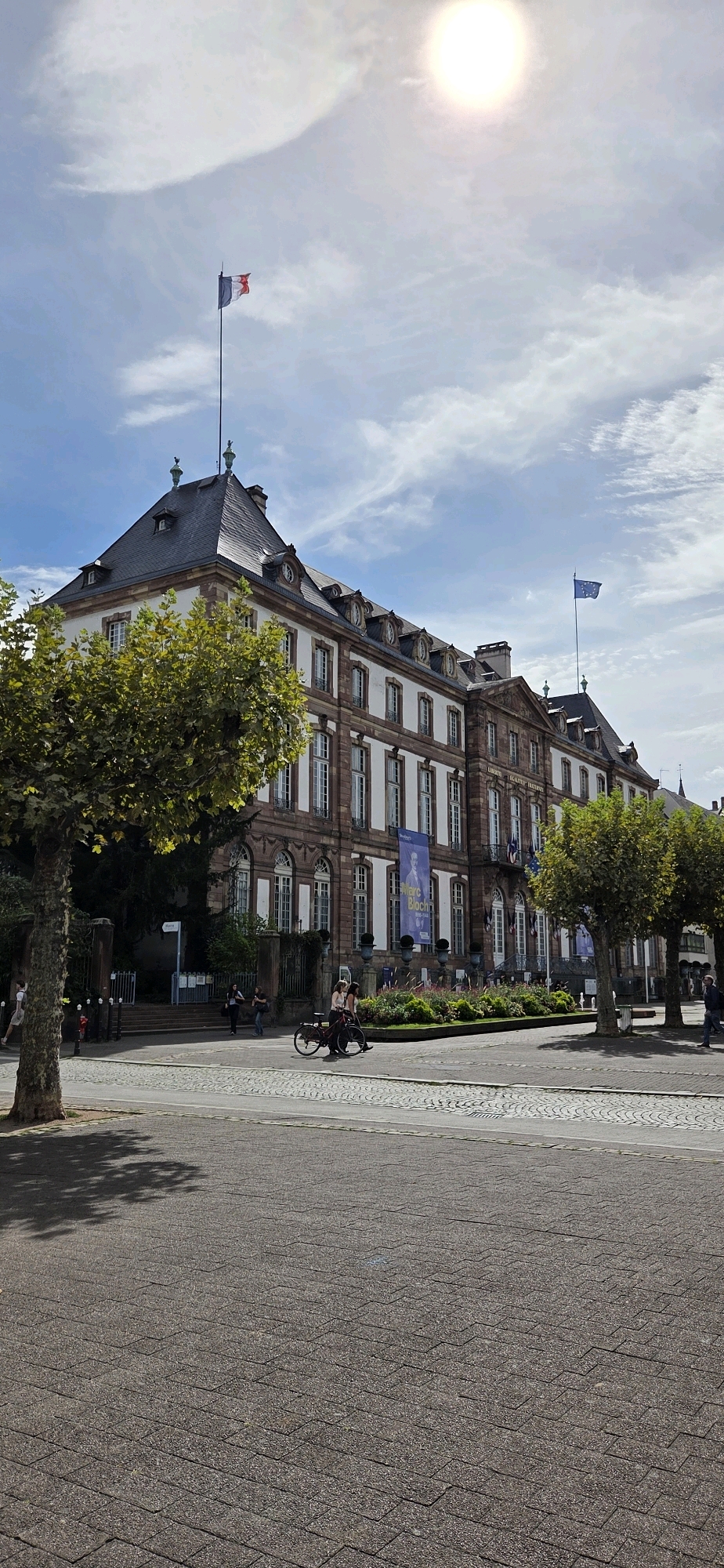 The image depicts a stately building with a mansard roof in Strasbourg, France, likely the Palais Rohan due to its architecture and the presence of flags. A French flag flies from a flagpole atop the building, and an EU flag is visible on another flagpole to the right. The building's facade is a mix of white and reddish-brown stone, with numerous arched windows and decorative elements.
In the foreground, a paved area transitions from rougher stone to smoother asphalt. A person is cycling on a red bicycle in the middle ground, with two passengers. Other individuals are walking on the sidewalk near the building. Lush green trees frame the scene on the left and right. The sky is partly cloudy with a bright sun, suggesting daytime. A large banner featuring a portrait and text is hung on the building, possibly advertising an exhibition. The environment appears to be a public square or a significant urban area.